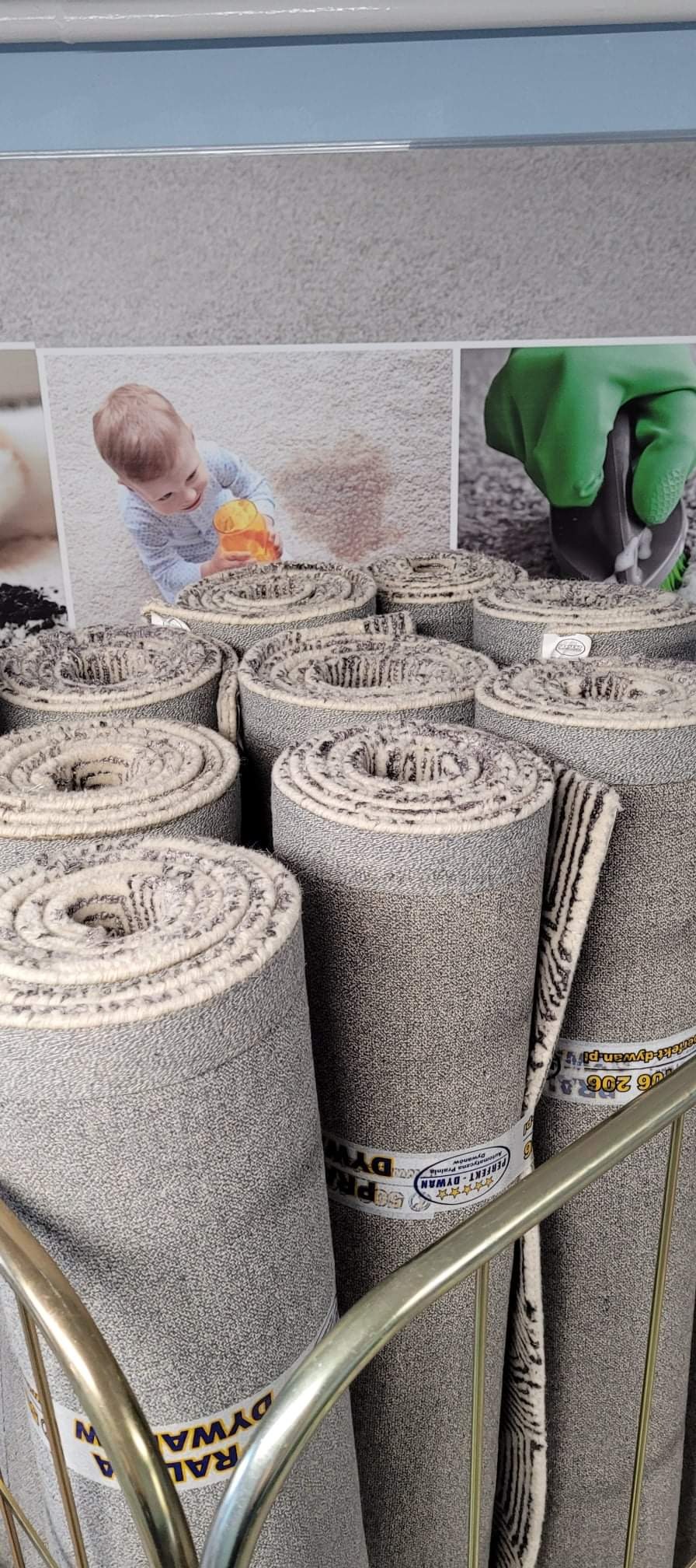 Stos zwiniętych dywanów w sklepie, na etykietach widoczne logo firmy 'Perfekt-Dywan', w tle zdjęcia przedstawiające dziecko bawiące się na dywanie i czyszczenie dywanu.