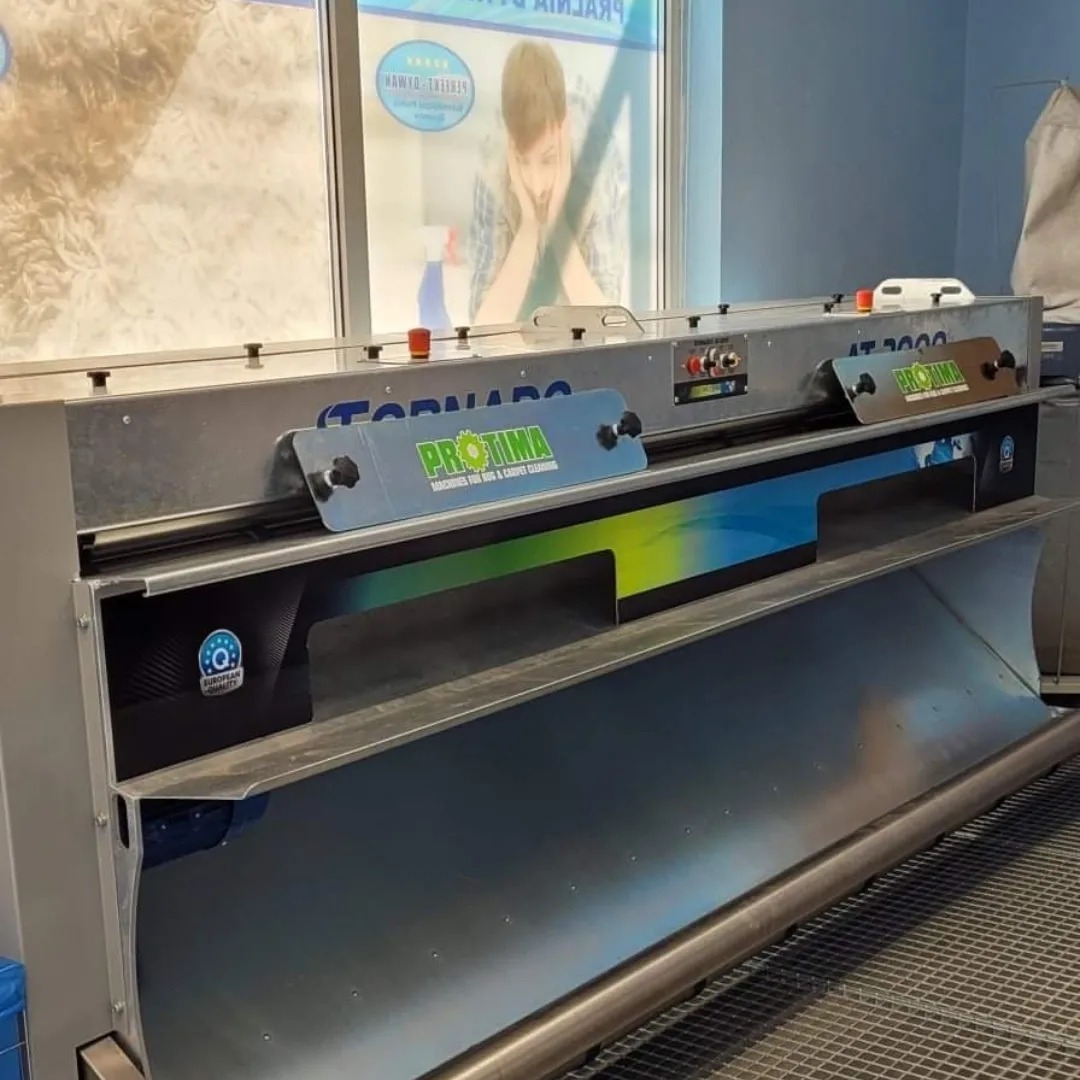 Profesjonalna maszyna do czyszczenia dywanów marki ProTima, model Tornado AT 2000, widoczna w pralni, z fragmentami dywanów w tle za szybą.