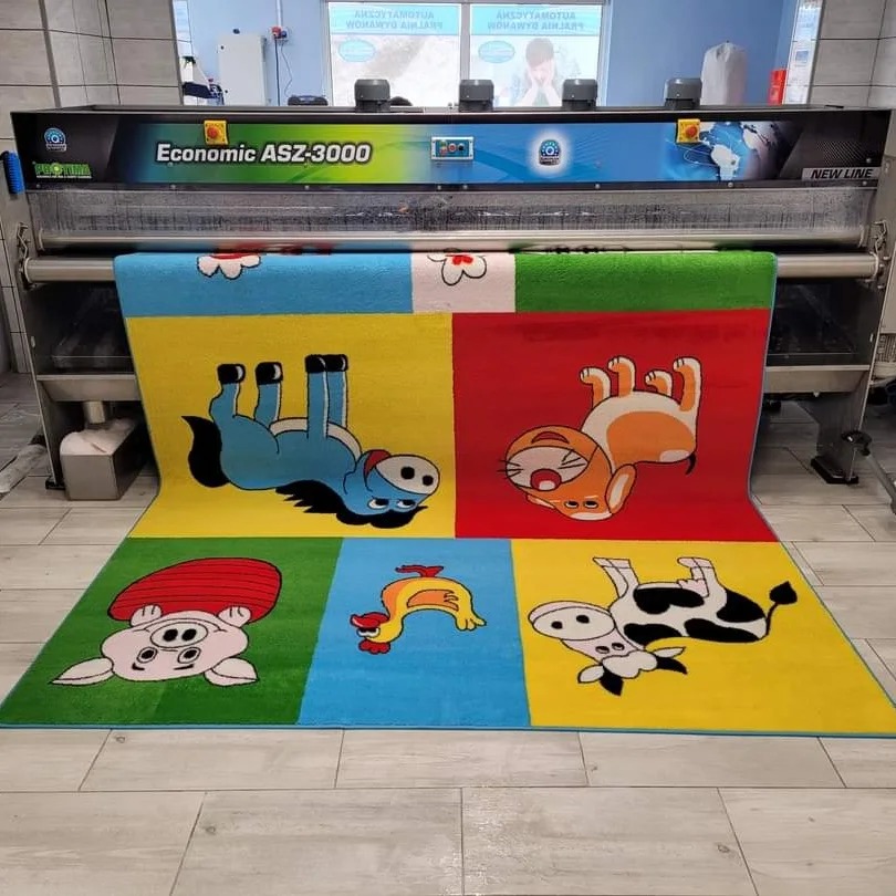 Wielobarwny dywan dziecięcy z motywami zwierząt przechodzi przez przemysłową maszynę czyszczącą Economic ASZ-3000 w pralni.