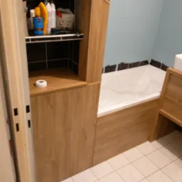 Łazienka z zabudową z płyty drewnopodobnej, obejmująca szafkę z półkami i obudowę wanny, biała wanna zabezpieczona folią, na półce kosmetyki, na podłodze białe płytki, widoczny nóż do tapet.