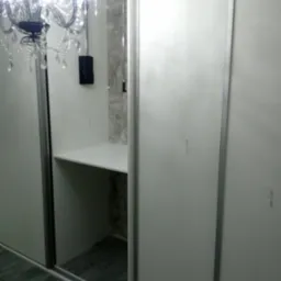 Biała szafa wnękowa z przesuwnymi drzwiami, widoczna półka w środku, oświetlona ozdobnym żyrandolem.