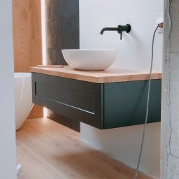 MebleRafi - Nowoczesna łazienka w trakcie wykańczania: biała umywalka nablatowa na drewnianym blacie z czarną szafką, widoczne fragmenty wanny i niedokończone obramowanie drzwi z przewodem elektrycznym.