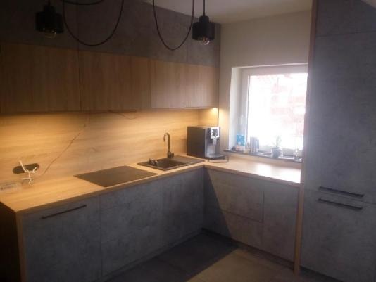 Nowoczesna kuchnia z szarymi frontami, drewnianym blatem i czarnymi lampami wiszącymi, widok na okno z roślinami doniczkowymi.