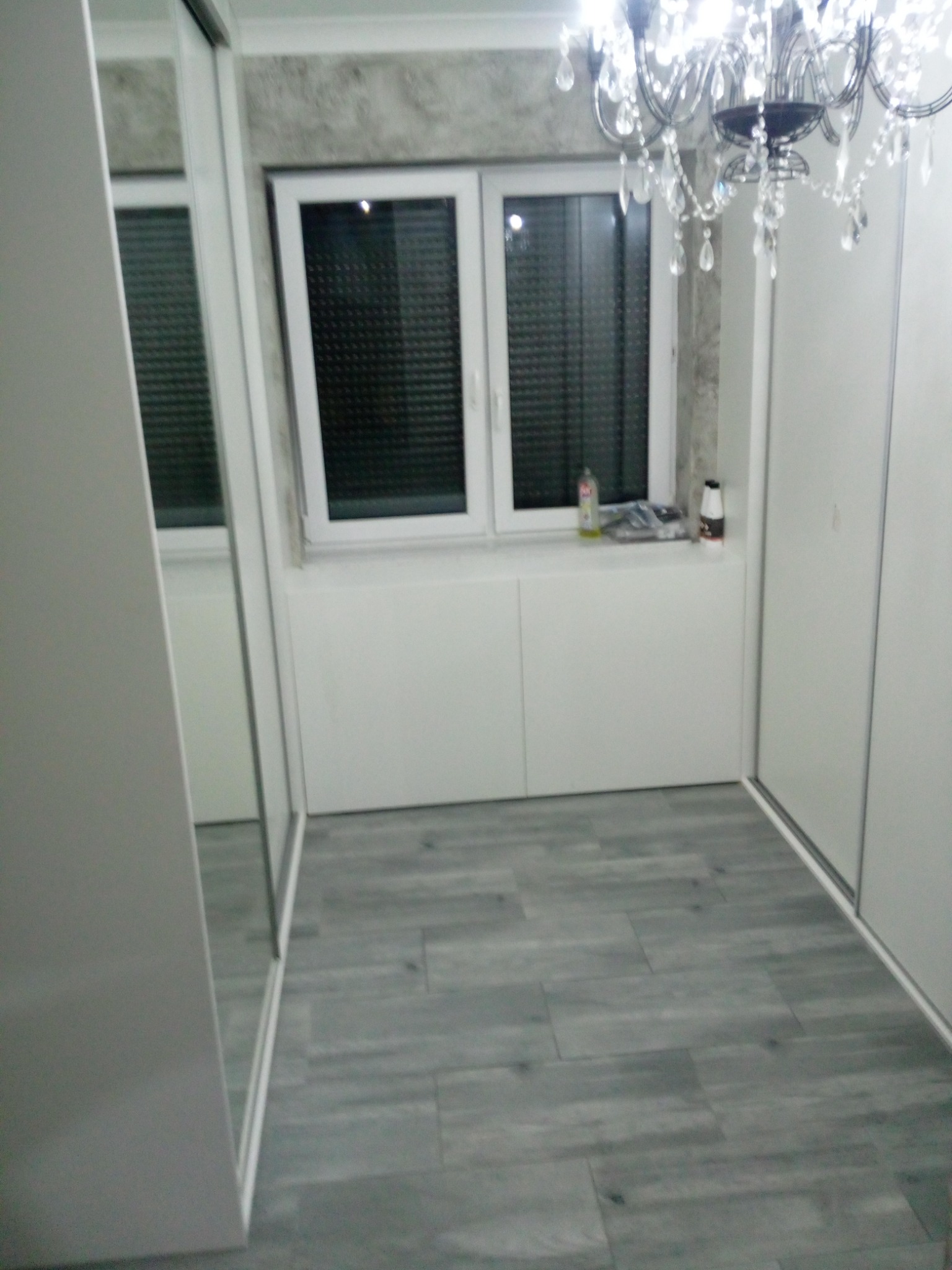 Wnętrze pokoju z szarą podłogą imitującą drewno, białą szafą przesuwną z lustrem po lewej, oknem z roletami i zabudową pod oknem, oraz ozdobnym żyrandolem kryształowym w górnej części kadru.