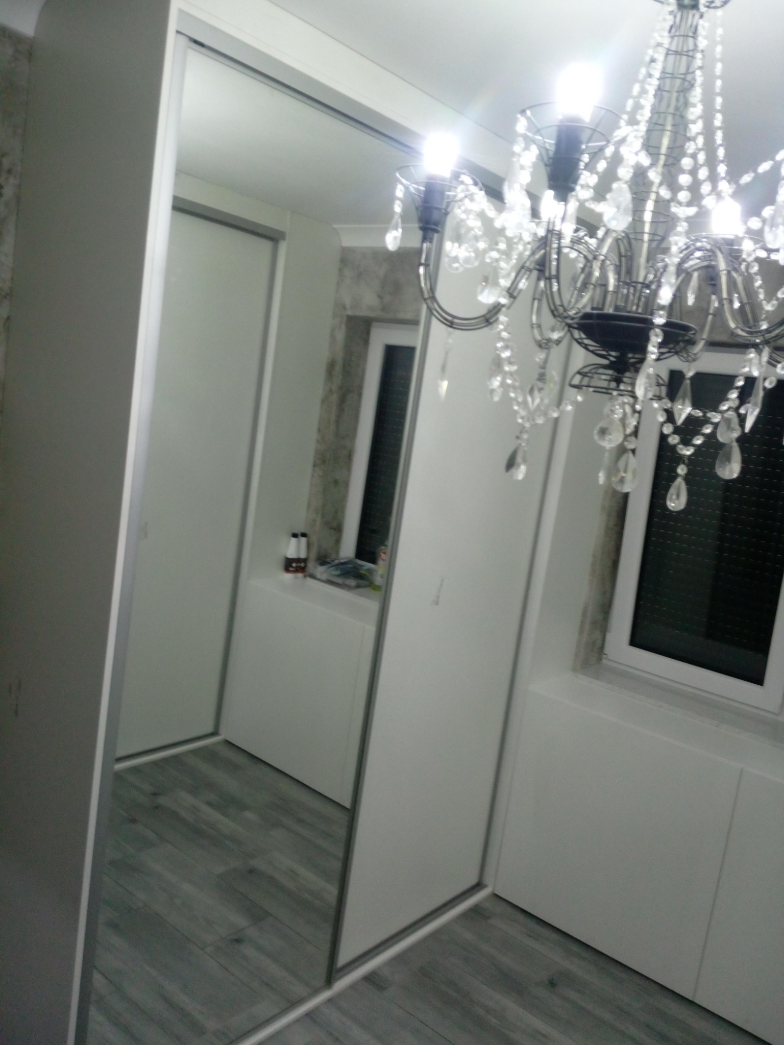 Szafa wnękowa z przesuwnymi, lustrzanymi drzwiami, odbijająca szary parkiet i żyrandol z kryształkami, w tle okno z żaluzjami.