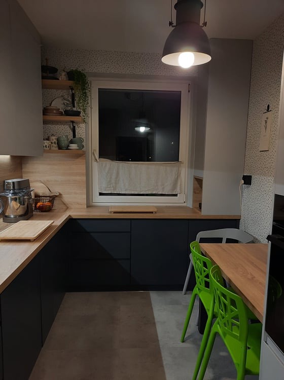 Nowoczesna kuchnia z ciemnymi, gładkimi frontami szafek, drewnianym blatem i oświetleniem punktowym, uzupełniona o zielone krzesła i drewniany stół.