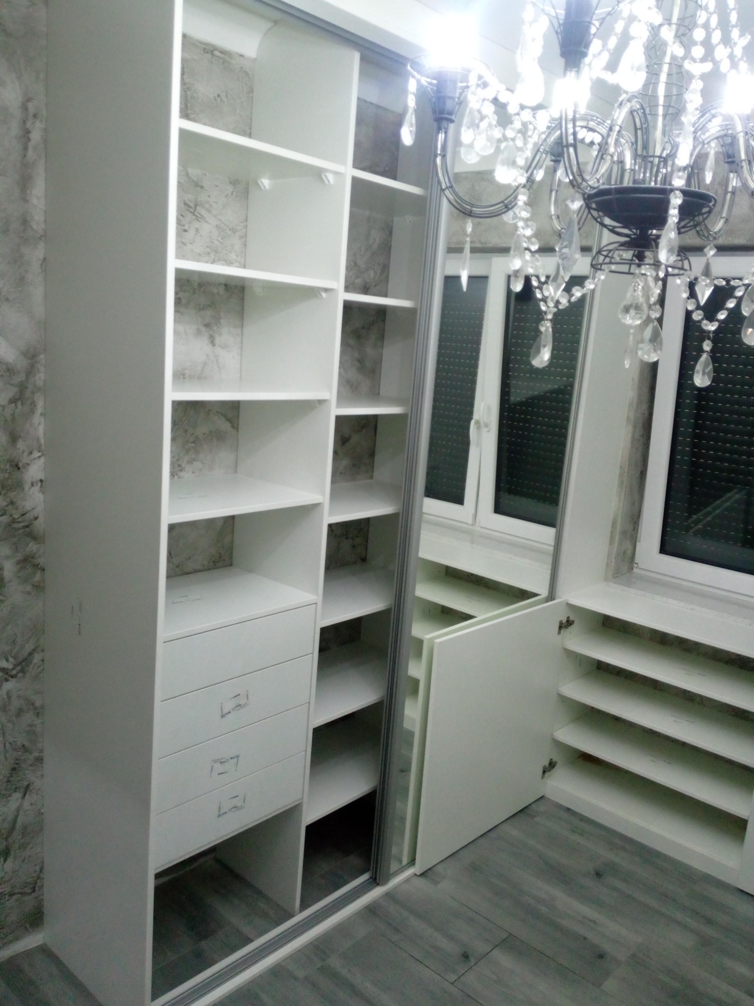 Biała szafa wnękowa z lustrzanymi drzwiami przesuwnymi, zlokalizowana w Żywcu, oświetlona przez ozdobny żyrandol, z widocznymi półkami i szufladami.