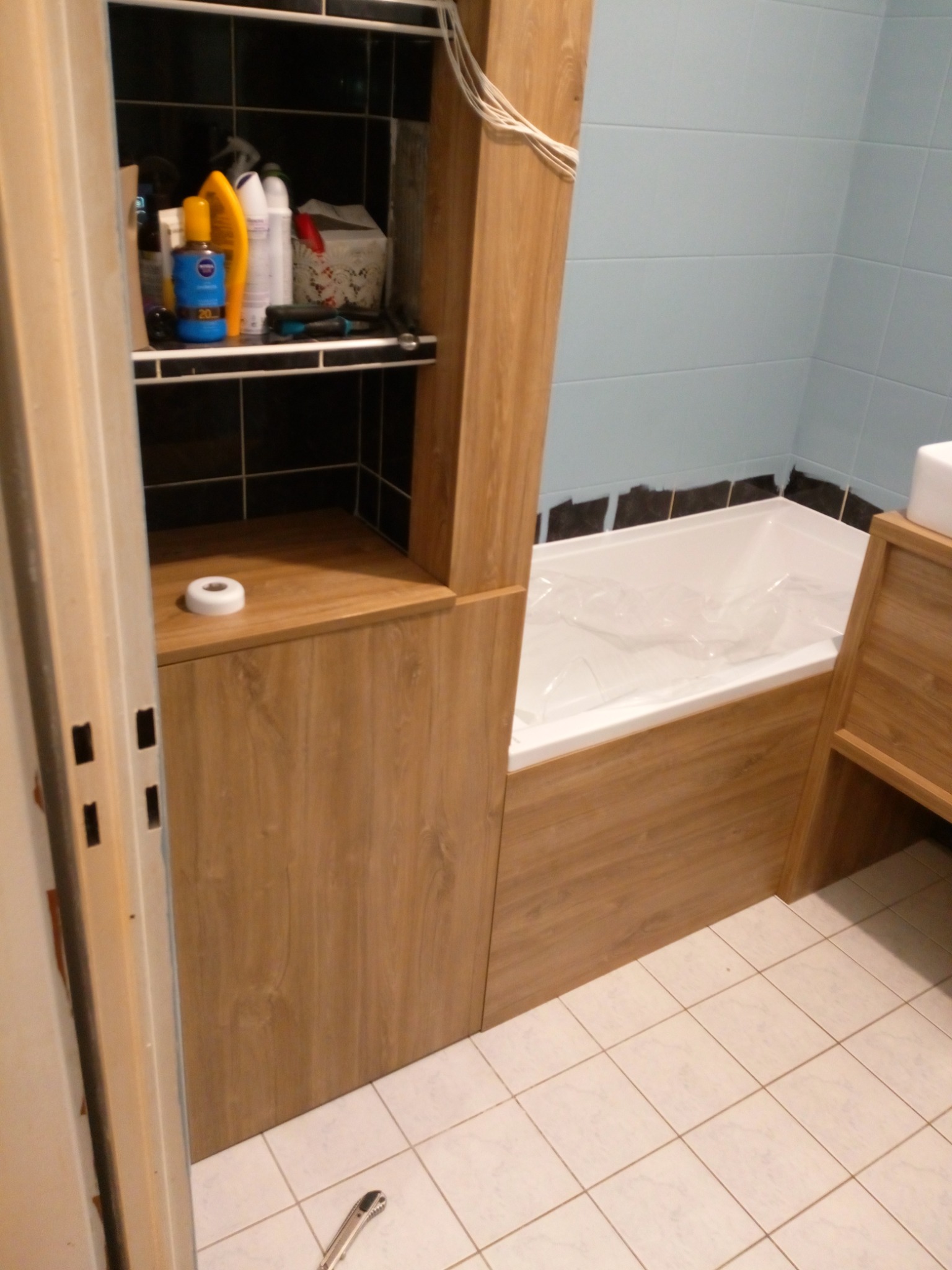 Łazienka z zabudową z płyty drewnopodobnej, obejmująca szafkę z półkami i obudowę wanny, biała wanna zabezpieczona folią, na półce kosmetyki, na podłodze białe płytki, widoczny nóż do tapet.
