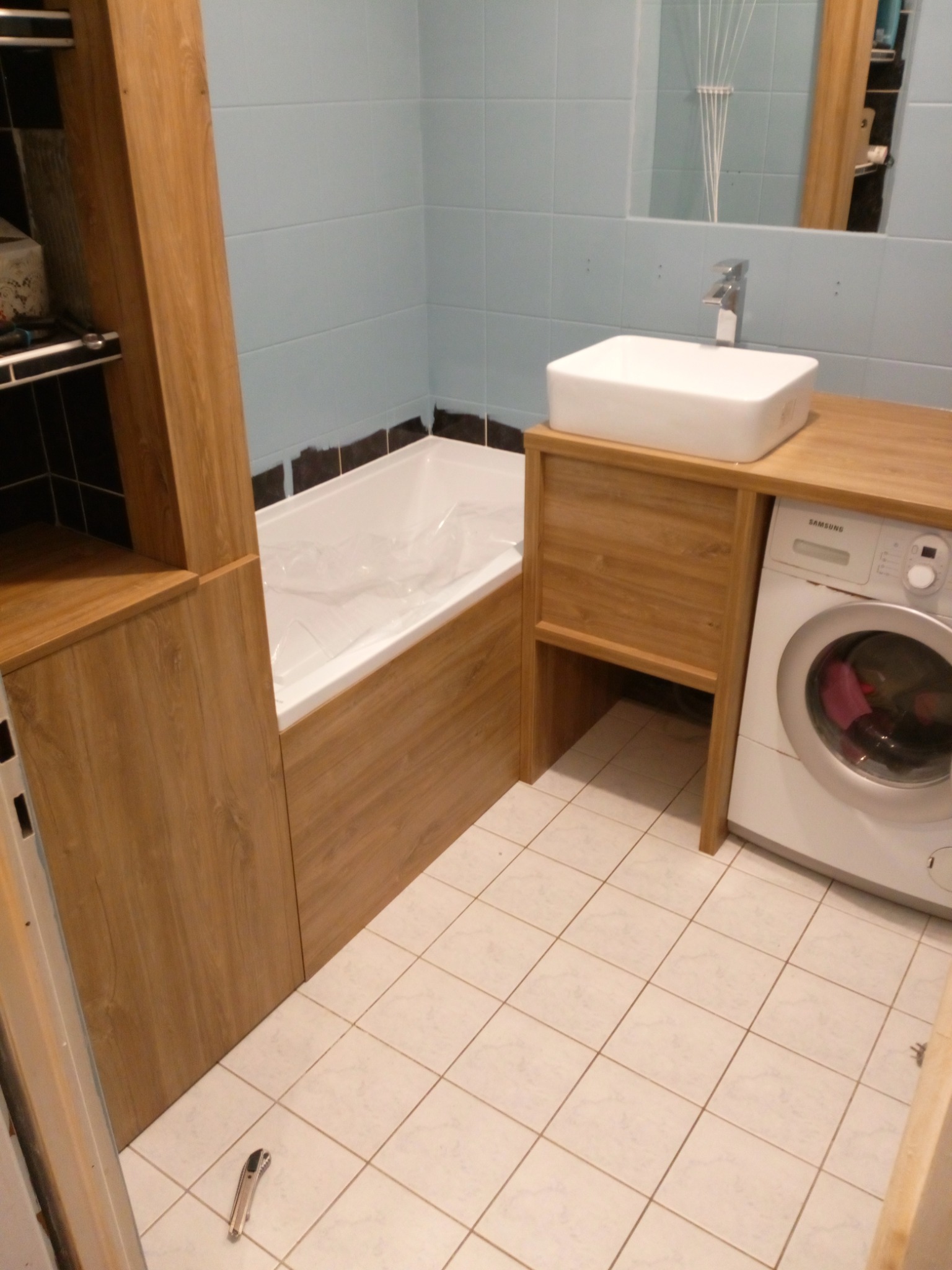 Łazienka z zabudową z drewnopodobnej płyty: szafka z umywalką, zabudowa wanny, słupek. Na podłodze leży nóż monterski.