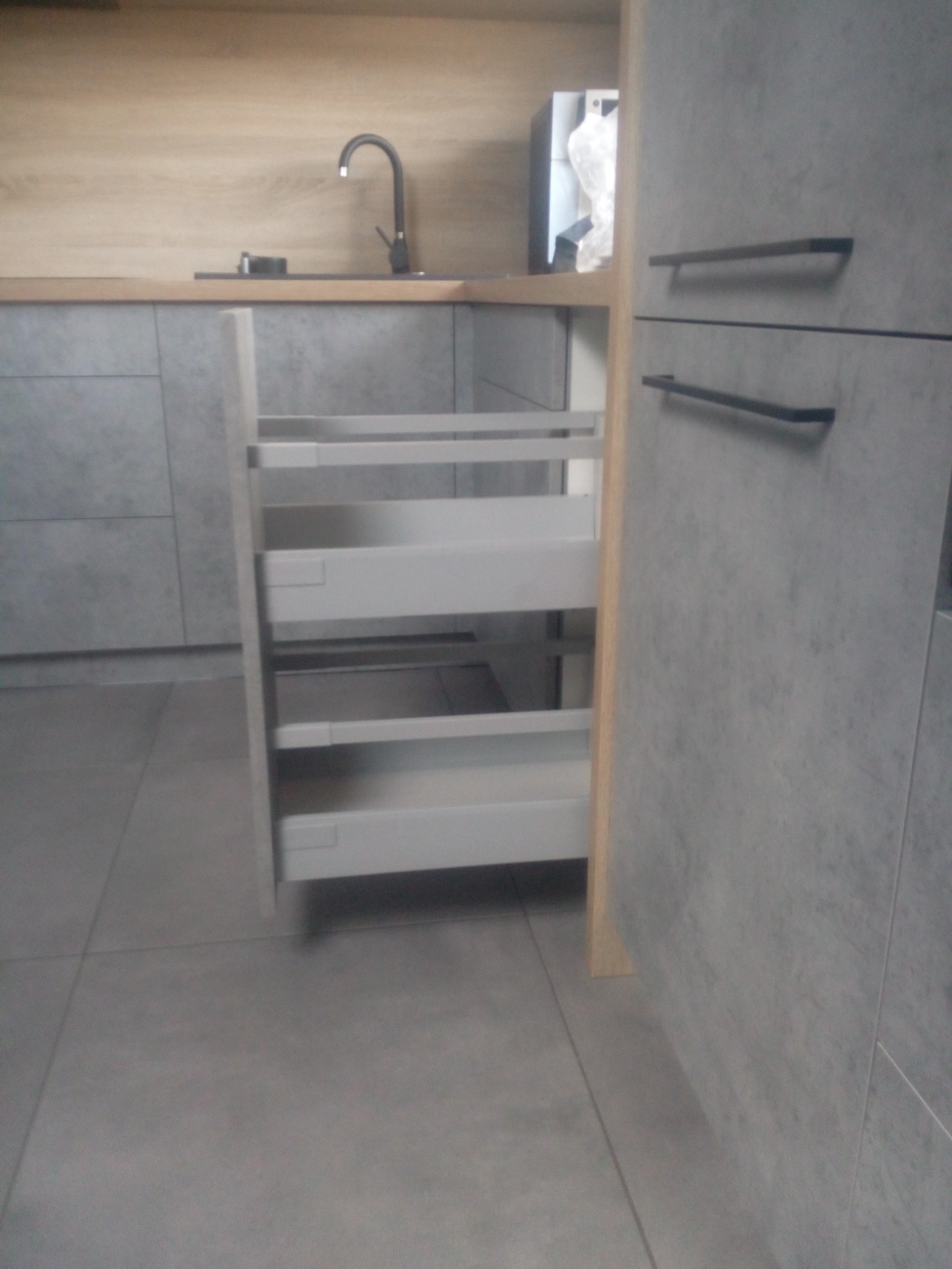 Szara kuchnia z wysuwanymi szufladami cargo, czarny kran i drewniany blat, widoczne płytki podłogowe.