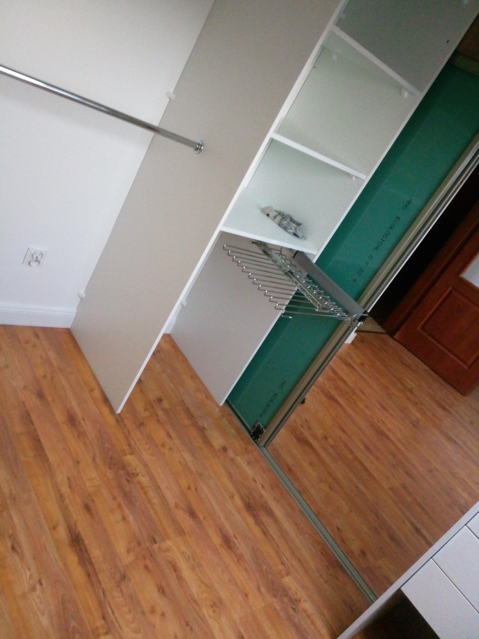 Wnętrze szafy przesuwnej z drążkiem na ubrania i półkami, widoczna na tle drewniana podłoga i fragment drzwi.