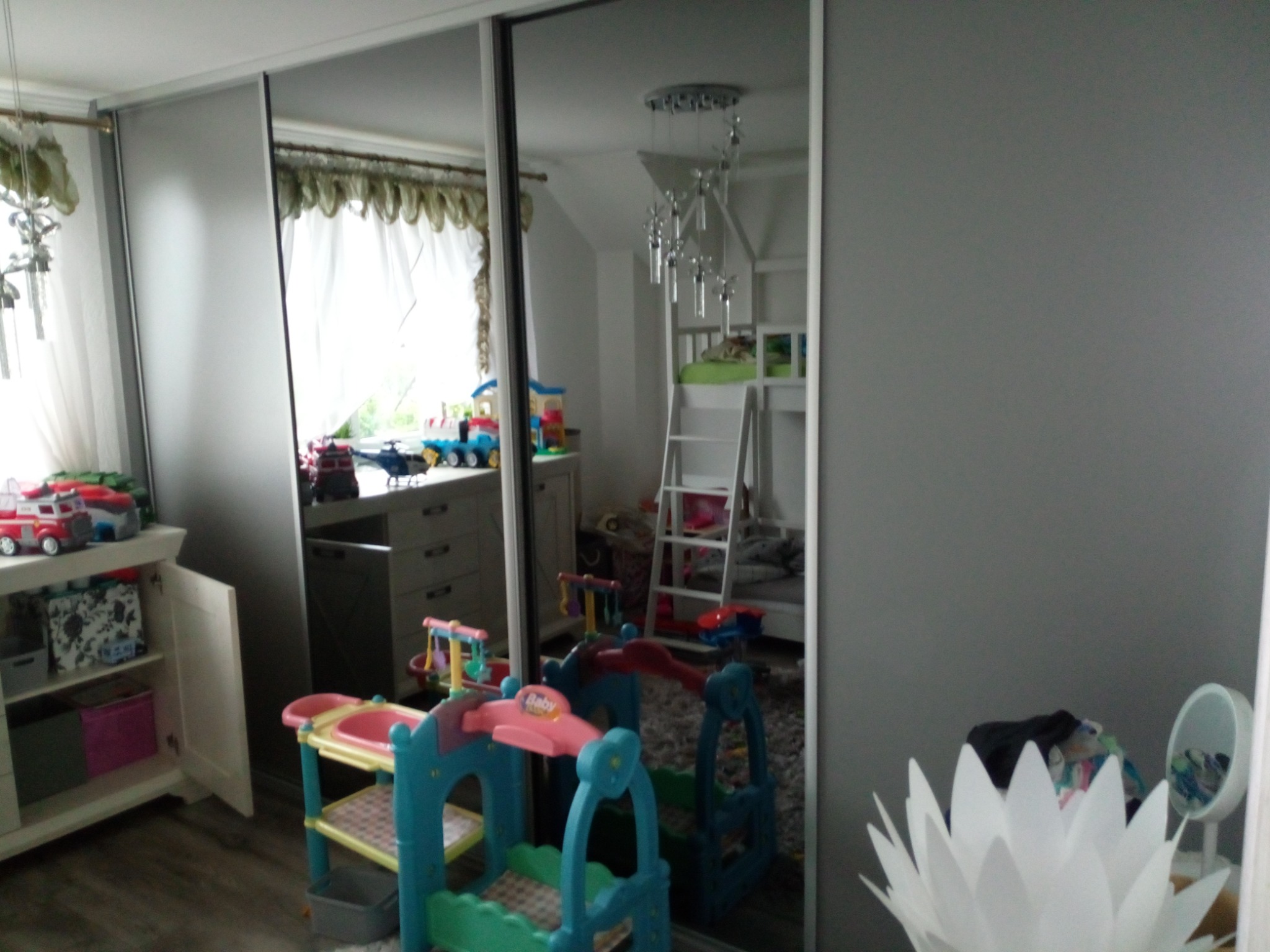 Szafa przesuwna z lustrzanymi drzwiami w pokoju dziecięcym, odbijająca łóżko piętrowe, zabawki i białą lampę w kształcie kwiatu.