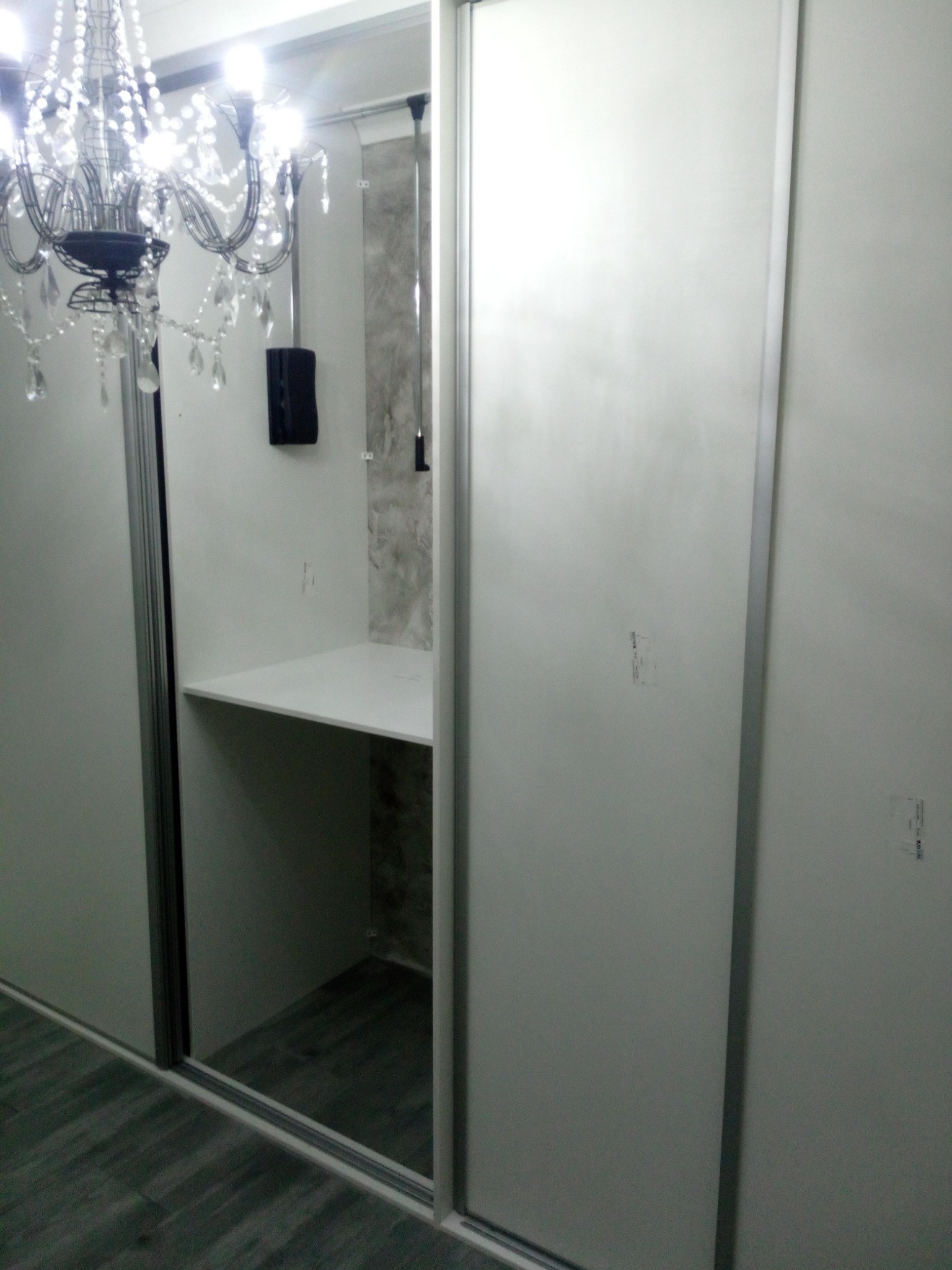 Biała szafa wnękowa z przesuwnymi drzwiami, widoczna półka w środku, oświetlona ozdobnym żyrandolem.