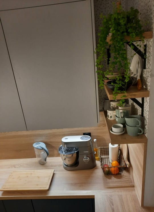 Szary robot kuchenny Kenwood na blacie z drewnopodobnym wzorem, obok deska do krojenia, w tle szafki kuchenne w odcieniach szarości i półki z roślinami oraz naczyniami.