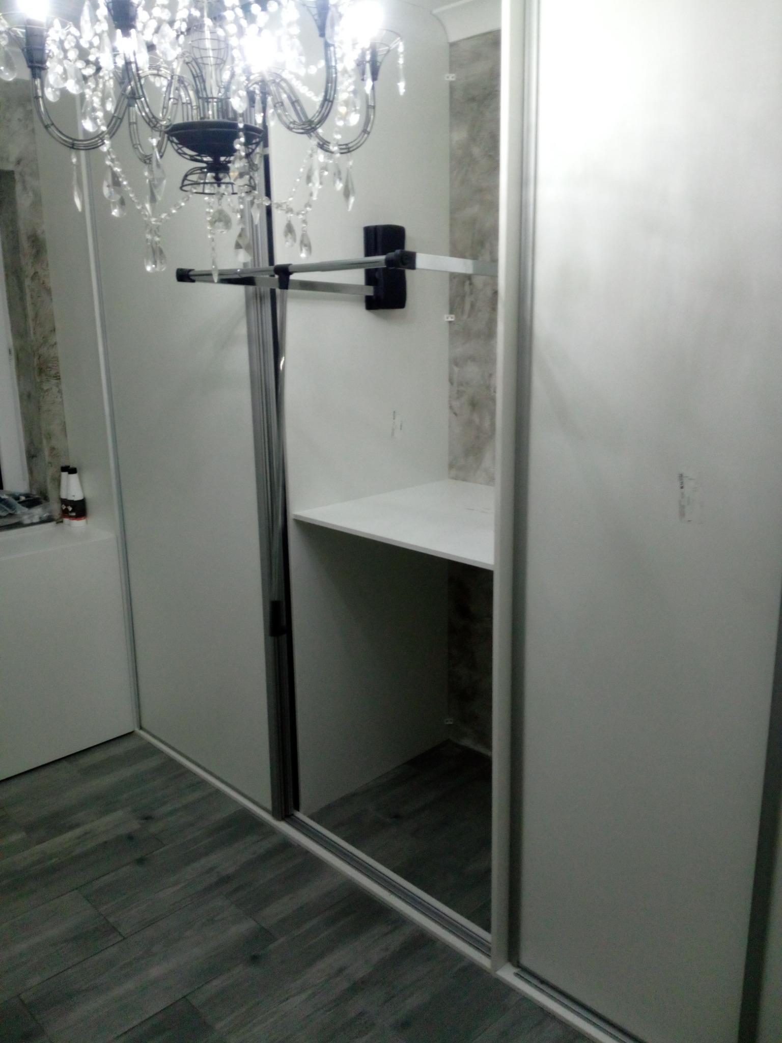 Wnętrze szafy przesuwnej z drążkiem na ubrania i półką, widoczna z boku z fragmentem żyrandola kryształowego i szarymi panelami podłogowymi.