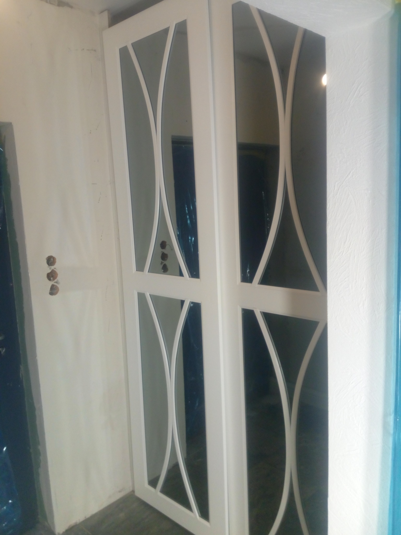 Biała szafa wnękowa z lustrami i dekoracyjnymi, zaokrąglonymi listwami, widoczne niedokończone prace wykończeniowe na ścianie.