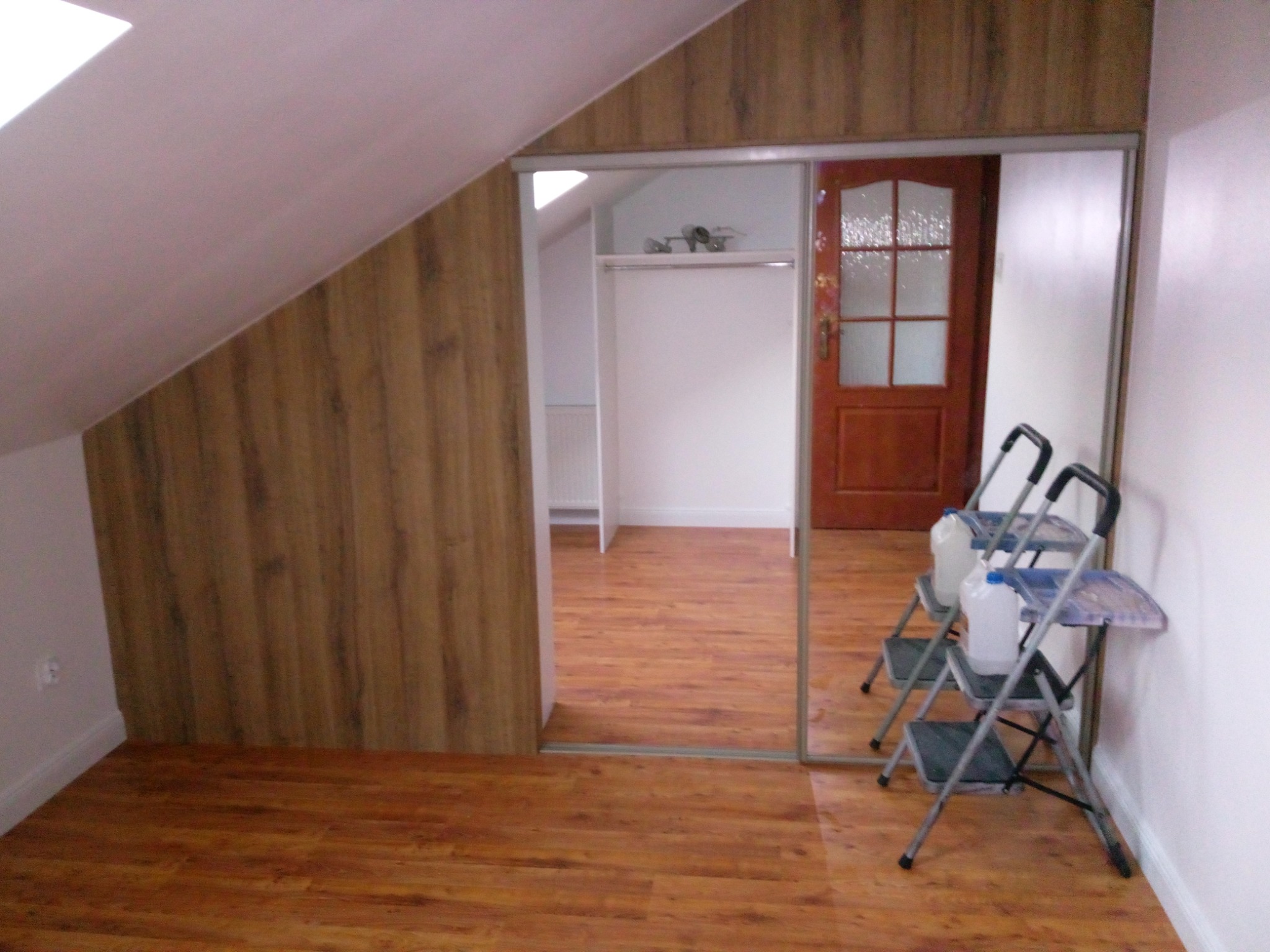 Poddasze z zamontowaną szafą wnękową z przesuwnymi lustrzanymi drzwiami, widoczne dwie drabiny malarskie i butelki z tworzywa sztucznego, drewniana podłoga i boazeria.