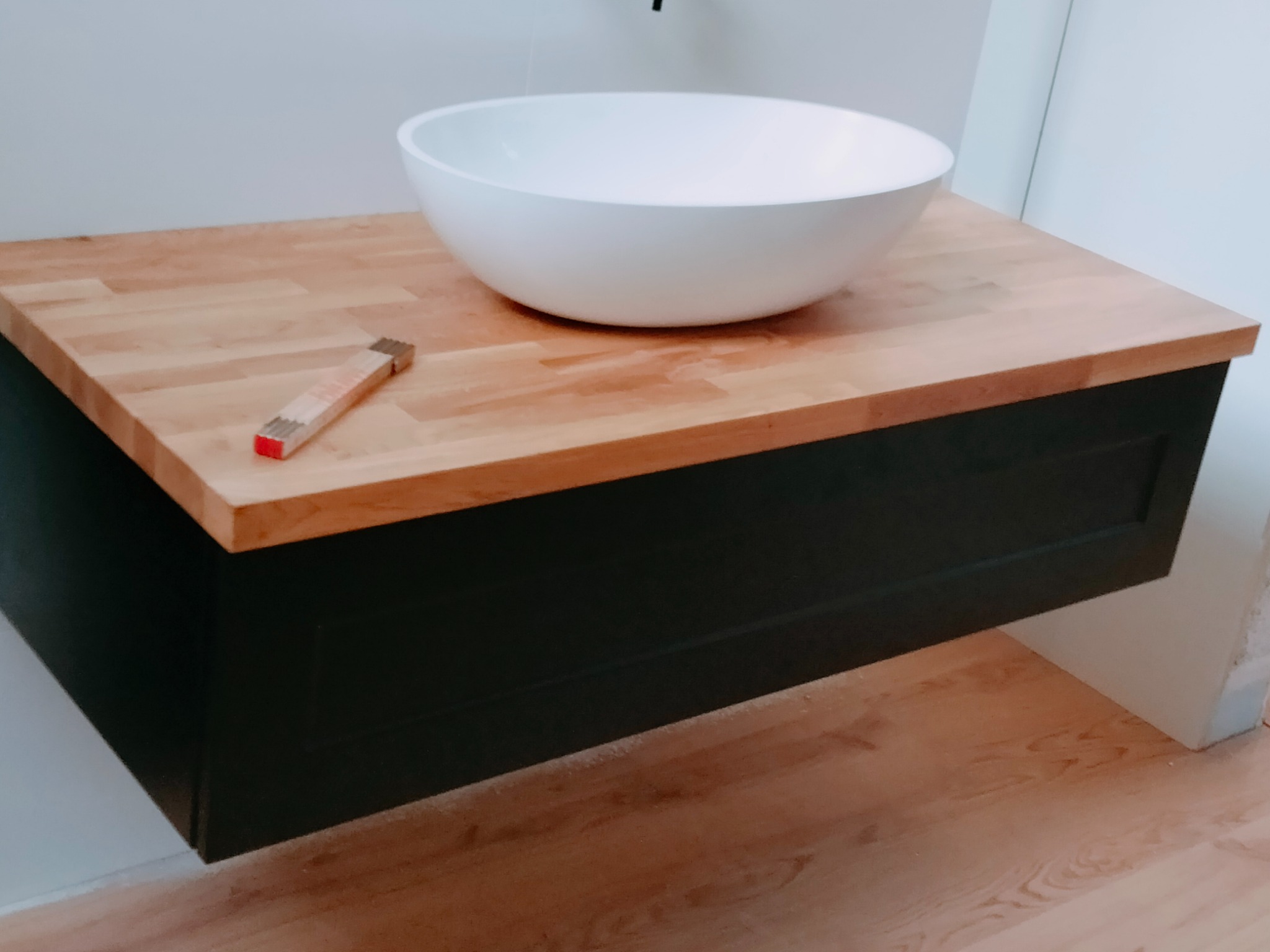 Biała umywalka nablatowa w kształcie misy na drewnianym blacie szafki łazienkowej w kolorze ciemnej zieleni, obok leży składana miarka.