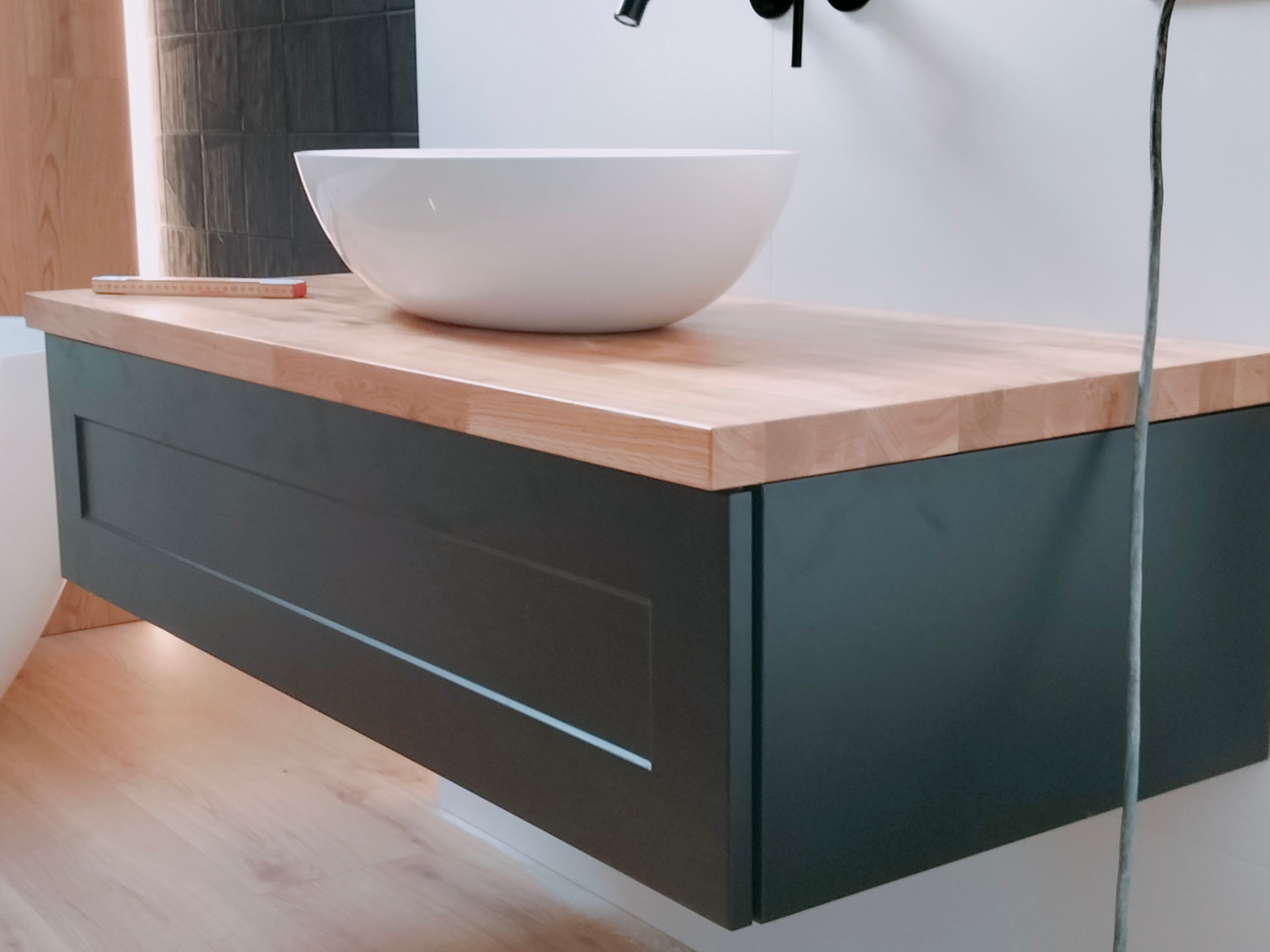 Nowoczesna łazienka z wiszącą szafką pod umywalkę z blatem z jasnego drewna, okrągła biała umywalka nablatowa, czarna bateria i linijka stolarska na blacie.