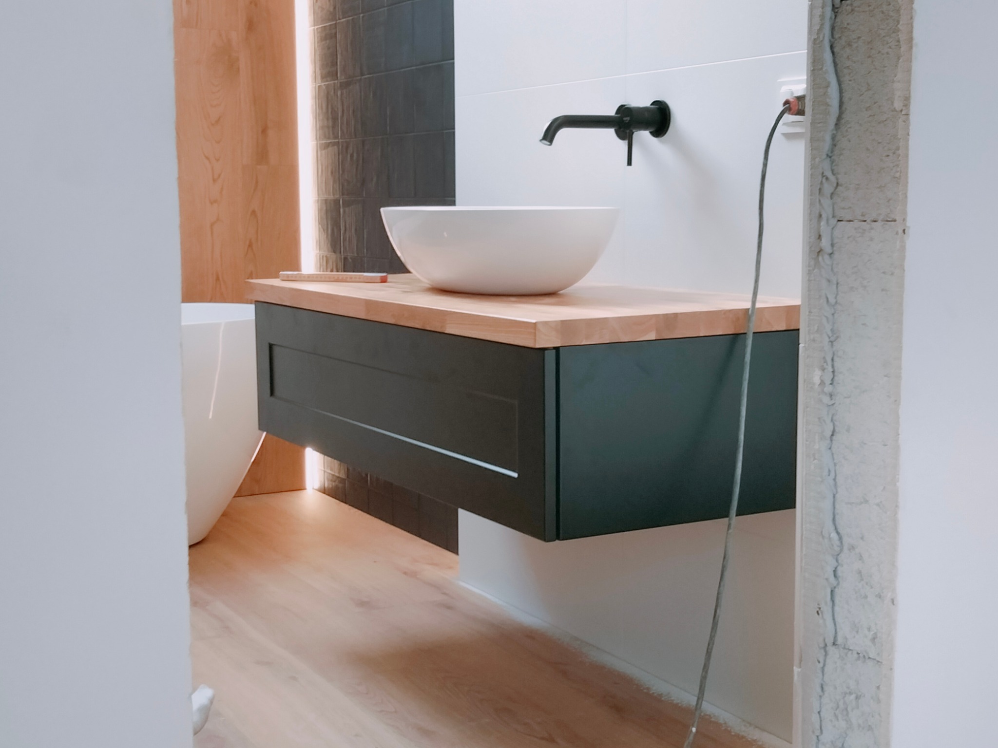 Nowoczesna łazienka w trakcie wykańczania: biała umywalka nablatowa na drewnianym blacie z czarną szafką, widoczne fragmenty wanny i niedokończone obramowanie drzwi z przewodem elektrycznym.