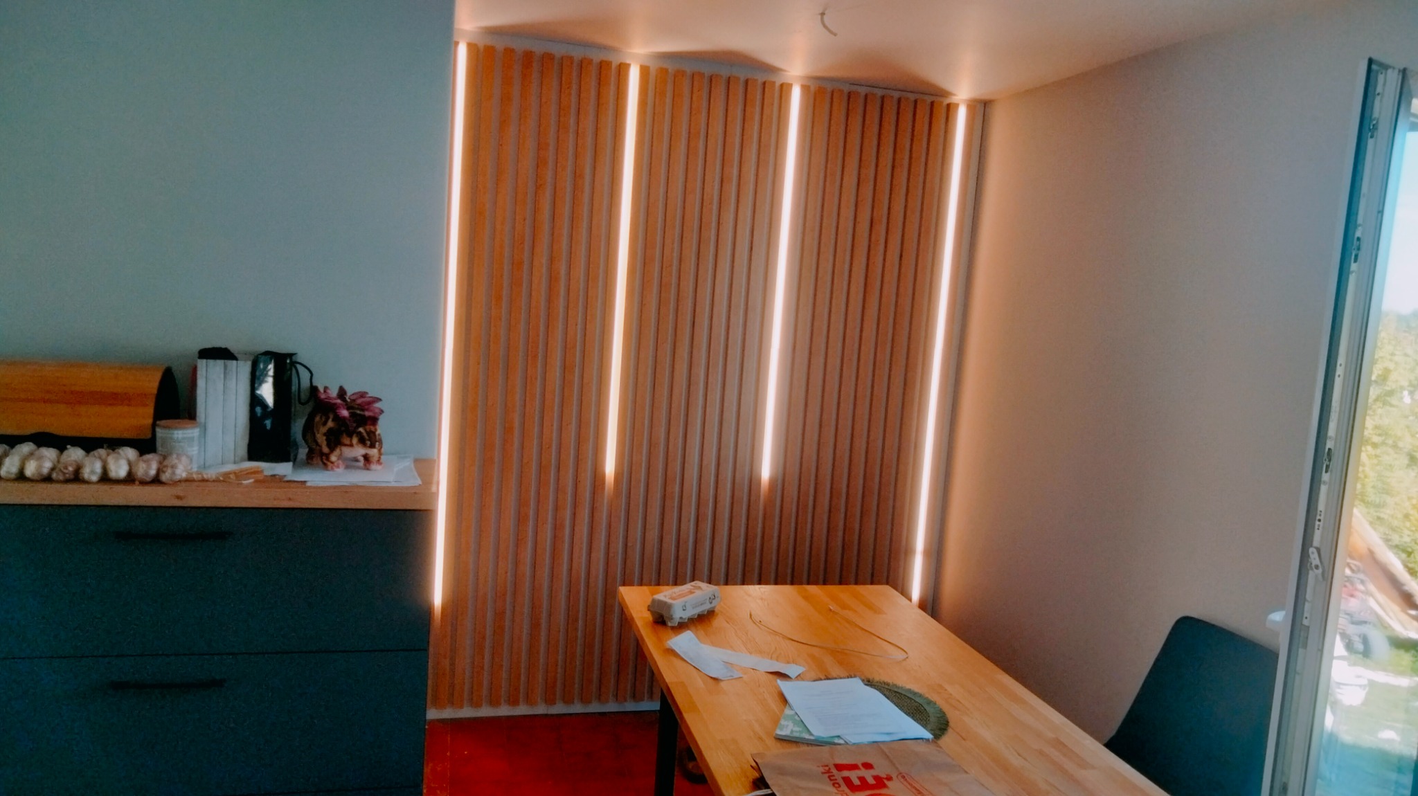 Nowoczesna kuchnia z drewnianymi lamelami podświetlonymi LED, blat z akcesoriami kuchennymi i dekoracjami, drewniany stół z dokumentami i torbą na zakupy w minimalistycznym wnętrzu.