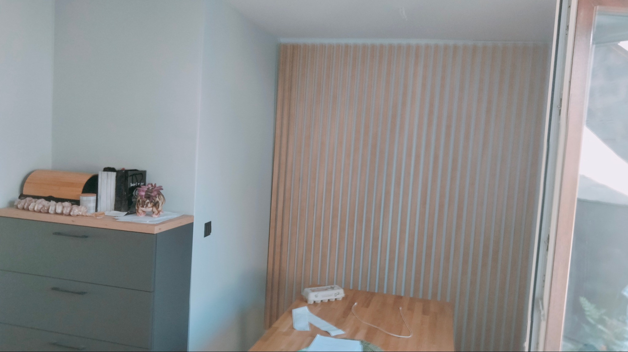 Panel ścienny z pionowych listew drewnianych, widok na fragment blatu stołu i szafkę z dekoracjami w tle.