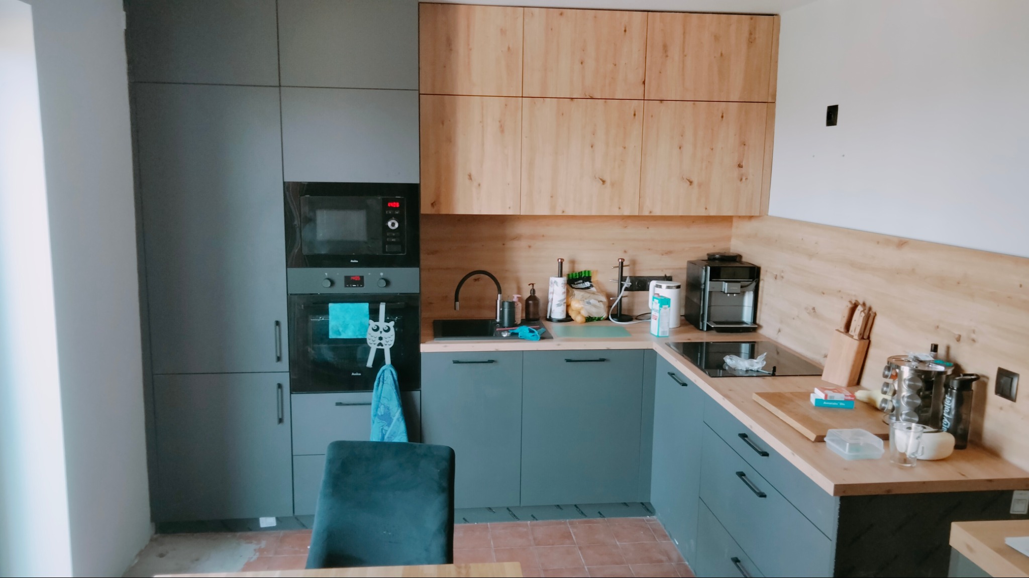 Nowoczesna kuchnia z szarymi i drewnopodobnymi frontami, czarnym piekarnikiem i płytą indukcyjną, widoczny ekspres do kawy i akcesoria kuchenne.