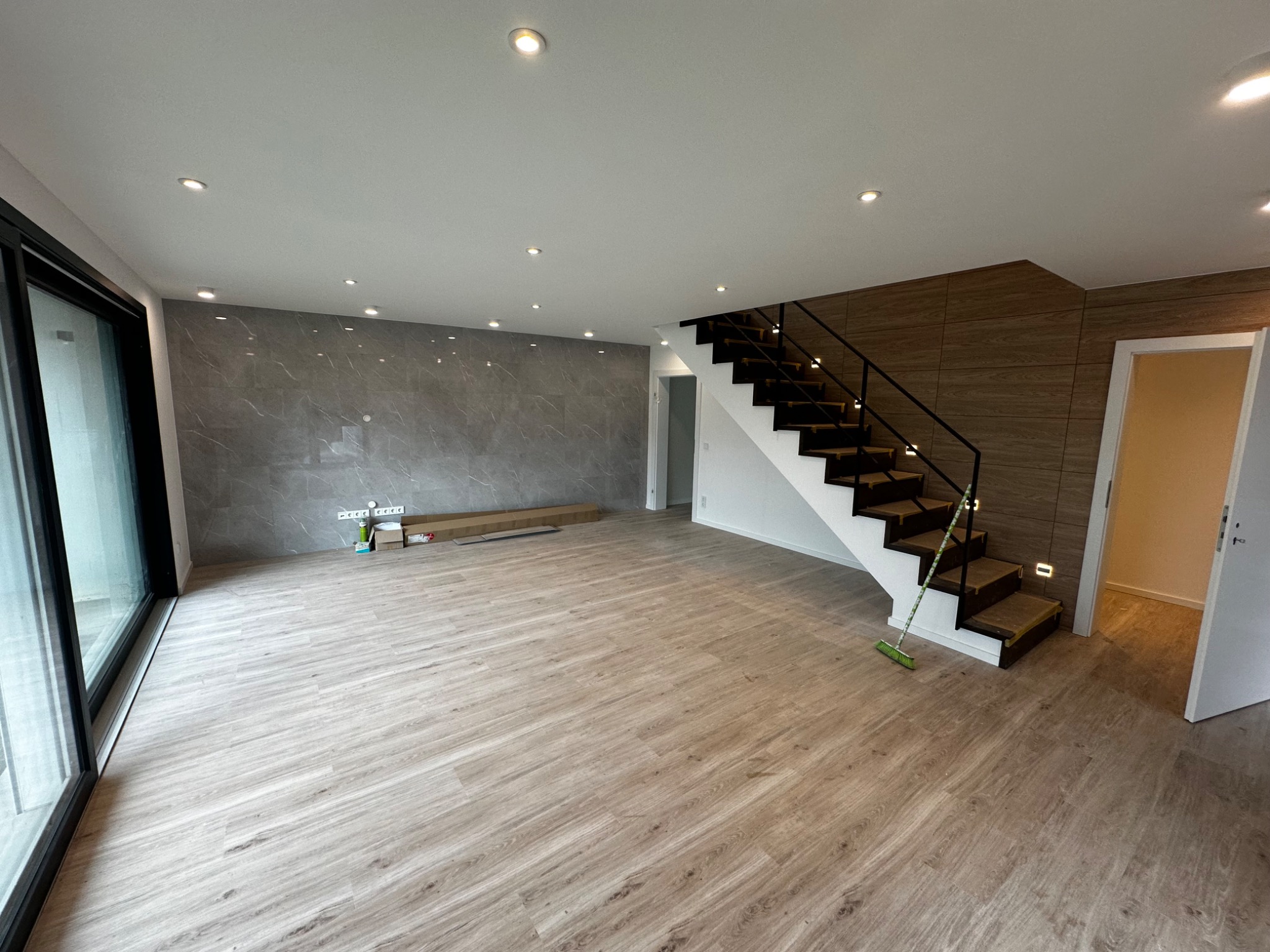 Przestronne wnętrze po remoncie z drewnopodobną podłogą, szarą ścianą z efektem marmuru i nowoczesnymi schodami z czarną balustradą, widoczne ślady po pracach wykończeniowych.