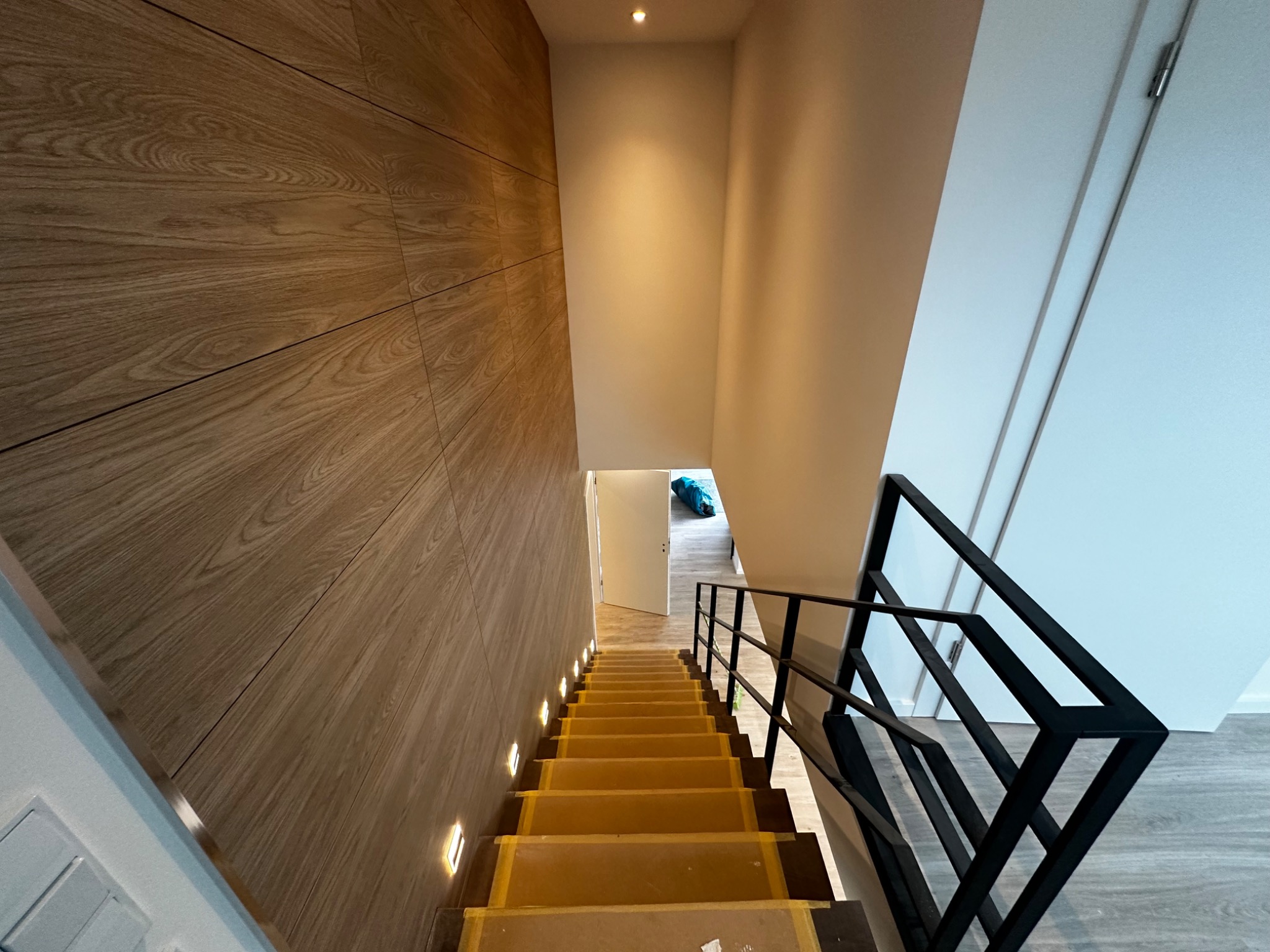 Schody wyłożone tymczasową folią ochronną, zabezpieczające stopnie podczas prac wykończeniowych; ściana wykończona panelami drewnopodobnymi z wbudowanym oświetleniem punktowym, czarna metalowa...