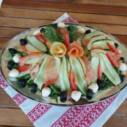 Srebrna taca z dekoracyjną kompozycją z łososia, ogórka, pomidora, czarnych oliwek, mini mozzarelli i koperku, ułożona na białej serwetce z czerwonym haftem, na drewnianym stole.