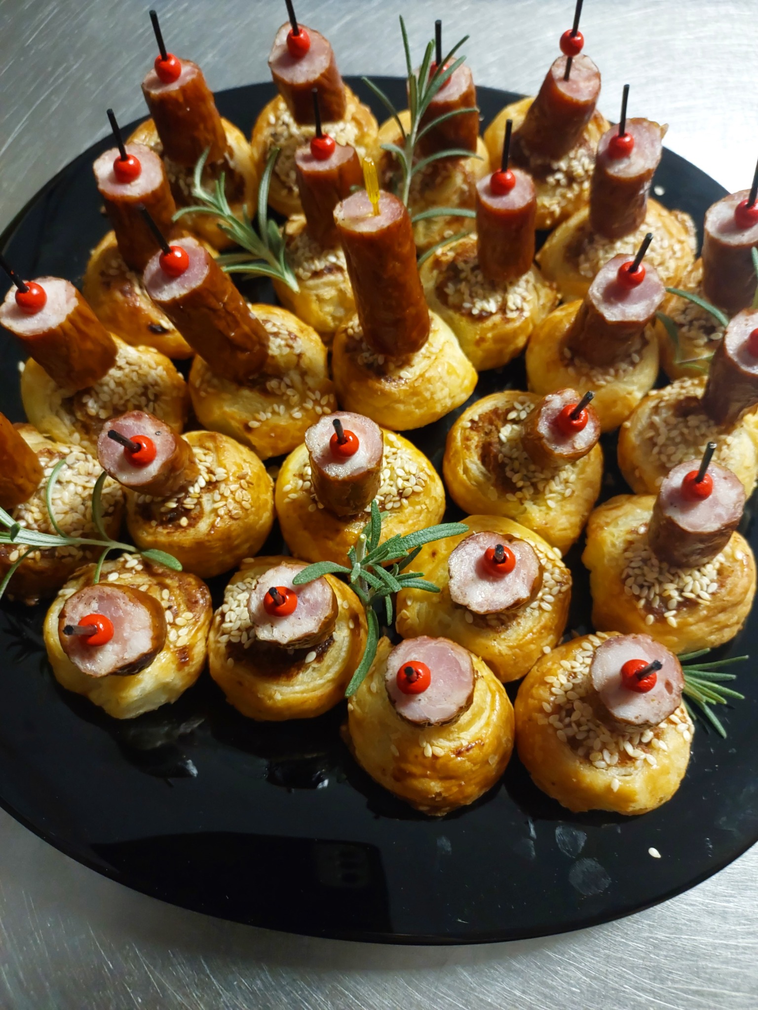 Miniaturowe przekąski: kruche babeczki posypane sezamem z plasterkiem kiełbasy i dekoracyjną czerwoną kulką na patyku, ułożone na czarnym talerzu, ozdobione gałązkami rozmarynu.