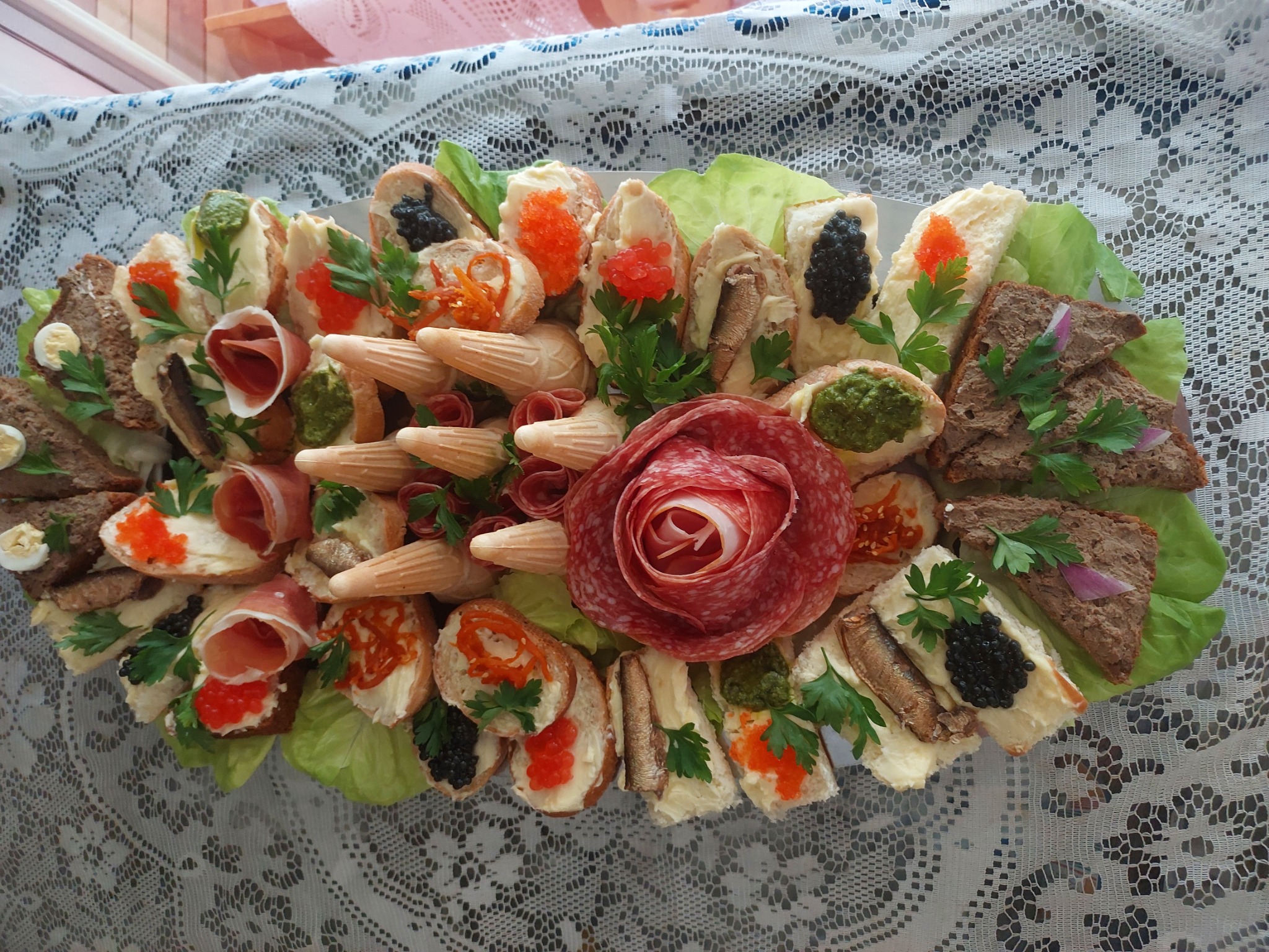 Dekoracyjna taca z kanapkami, przekąskami i wędlinami, ułożona na białym, koronkowym obrusie.