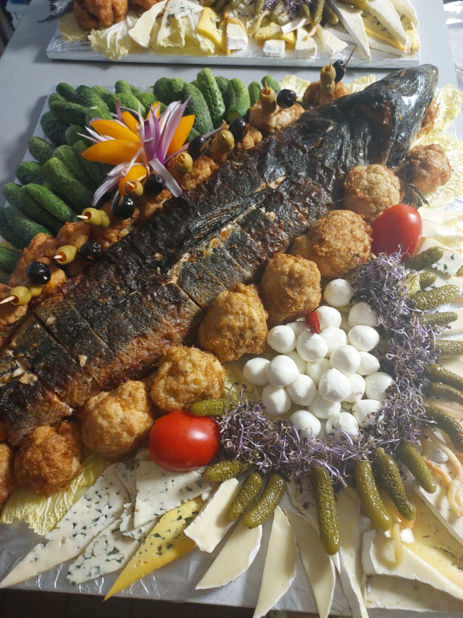 Dekoracyjna taca z pieczoną rybą, kulkami mięsnymi, serem pleśniowym, mini-mozzarellą, ogórkami kiszonymi i pomidorami, ozdobiona jadalnymi kwiatami i ziołami.