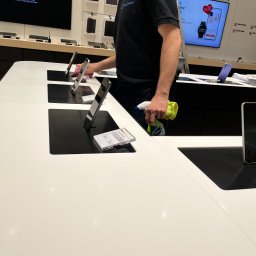 Clean Corner - Mateusz Zalewski - Mężczyzna w czarnej koszulce z logo firmy sprzątającej czyści ekspozycję smartfonów w sklepie, używając spryskiwacza Sidolux i żółtej ściereczki z mikrofibry.