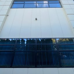 Clean Corner - Mateusz Zalewski - Wysoki budynek biurowy z nowoczesną elewacją w jasnych kolorach i oknami z ciemnymi ramami, w których odbija się otoczenie.