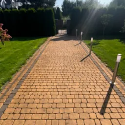 Żółta kostka brukowa prowadząca do bramy, oświetlona niskimi lampami ogrodowymi, otoczona zielonym trawnikiem i tujami, widok w słoneczny dzień.