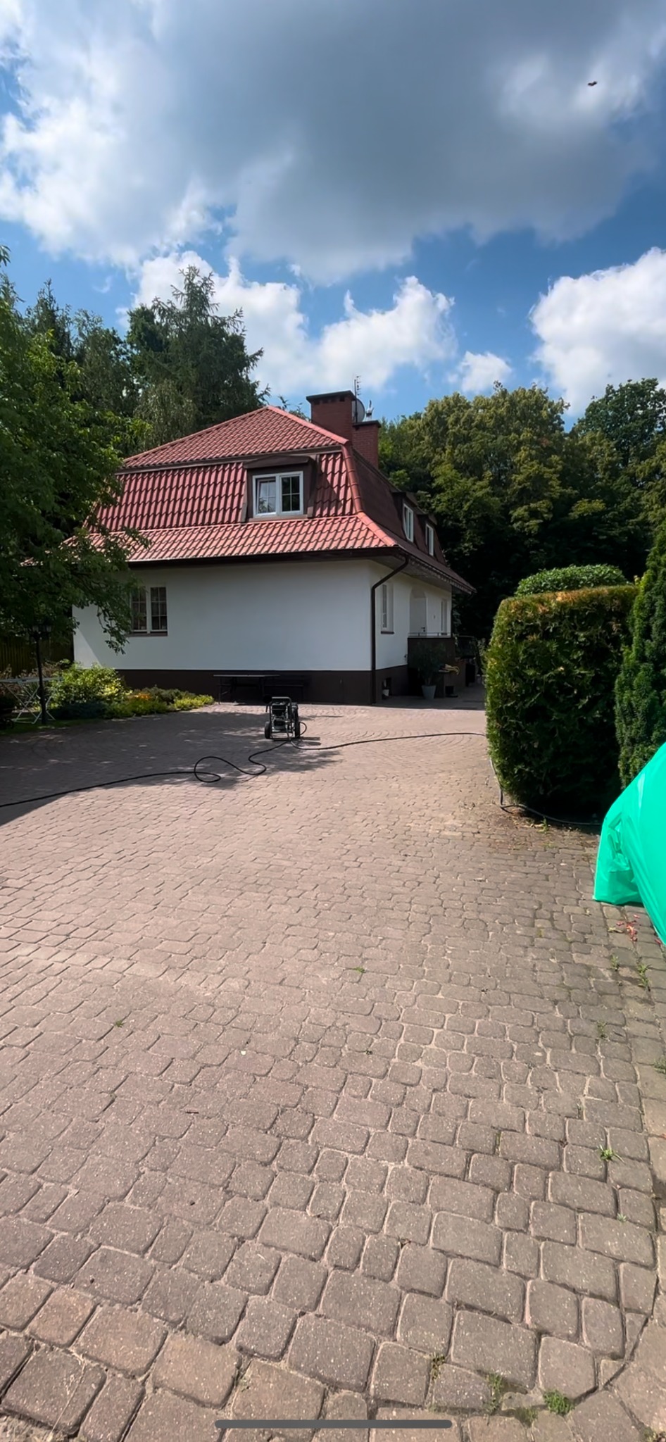 Dwupiętrowy dom z czerwonym dachem i białą elewacją, otoczony zielenią, z kostką brukową na podjeździe i myjką ciśnieniową gotową do użycia.
