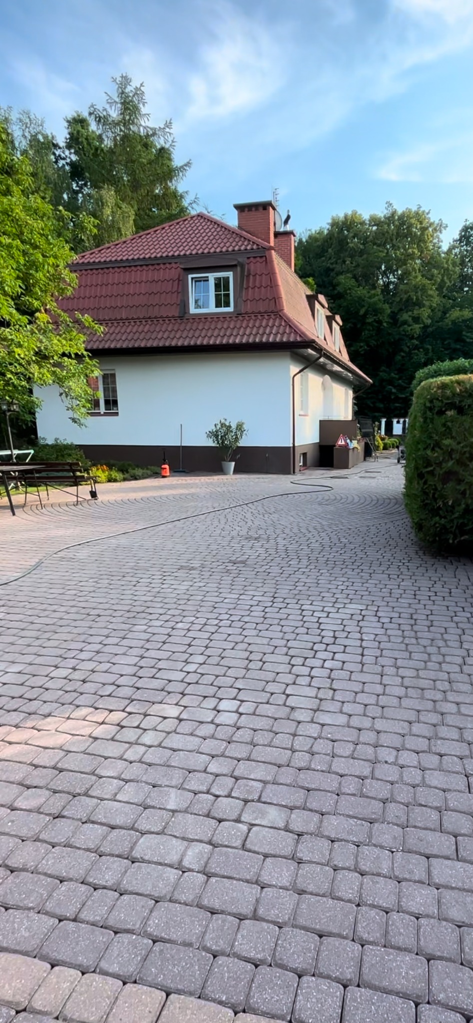 Szeroki kadr na podjazd z kostki brukowej prowadzący do białego domu z czerwoną dachówką, otoczonego zielenią.