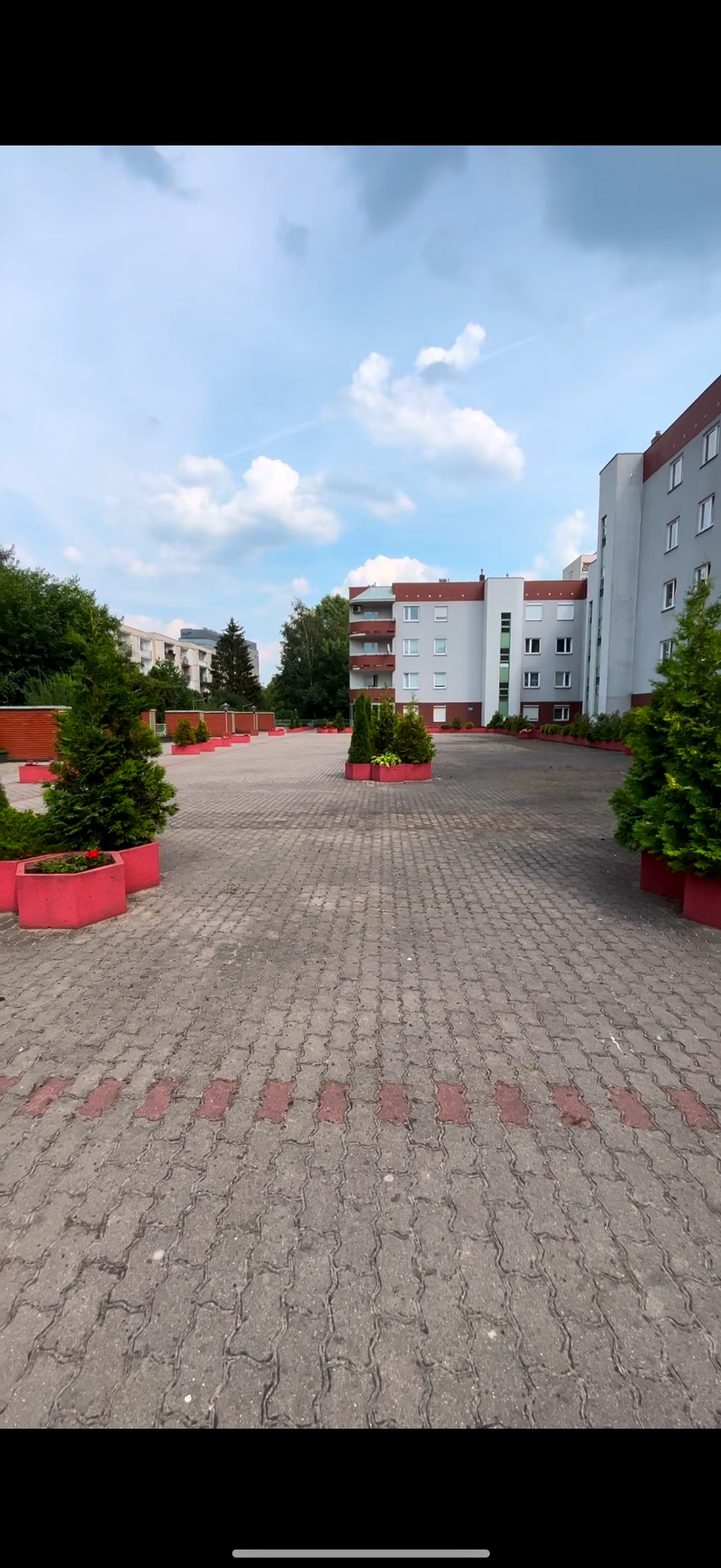 Kostka brukowa na parkingu przed budynkiem mieszkalnym, z czerwonymi akcentami wzdłuż krawędzi i w donicach z zielenią.