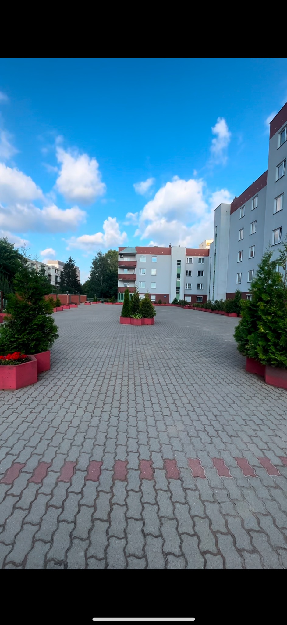 Szeroki dziedziniec wyłożony szarą kostką brukową z czerwonymi akcentami, otoczony budynkami mieszkalnymi i ozdobnymi rabatami z zielenią, fotografowany w słoneczny dzień z błękitnym niebem...