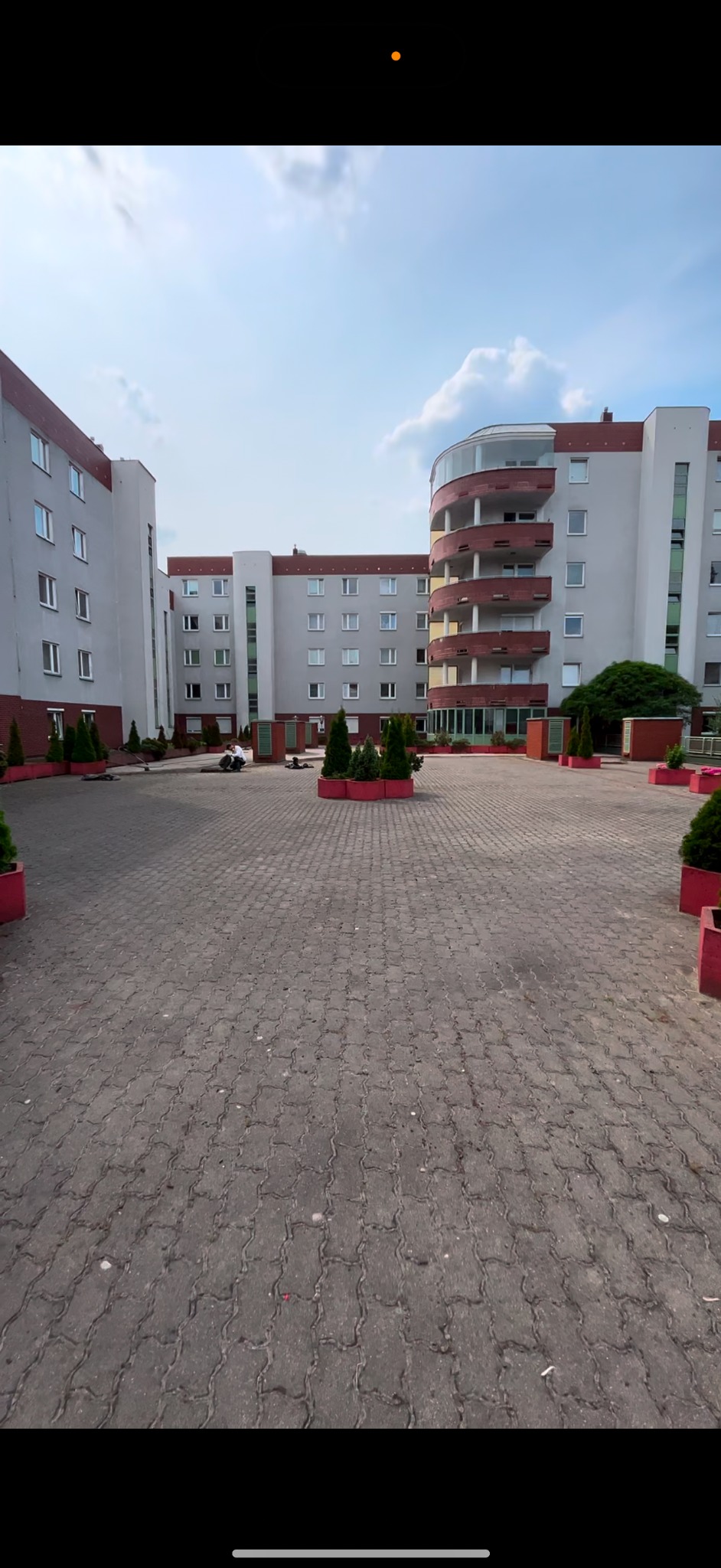 Dziedziniec osiedla mieszkaniowego z kostki brukowej, otoczony budynkami z szarymi elewacjami i czerwonymi akcentami, ozdobiony zielenią w donicach.