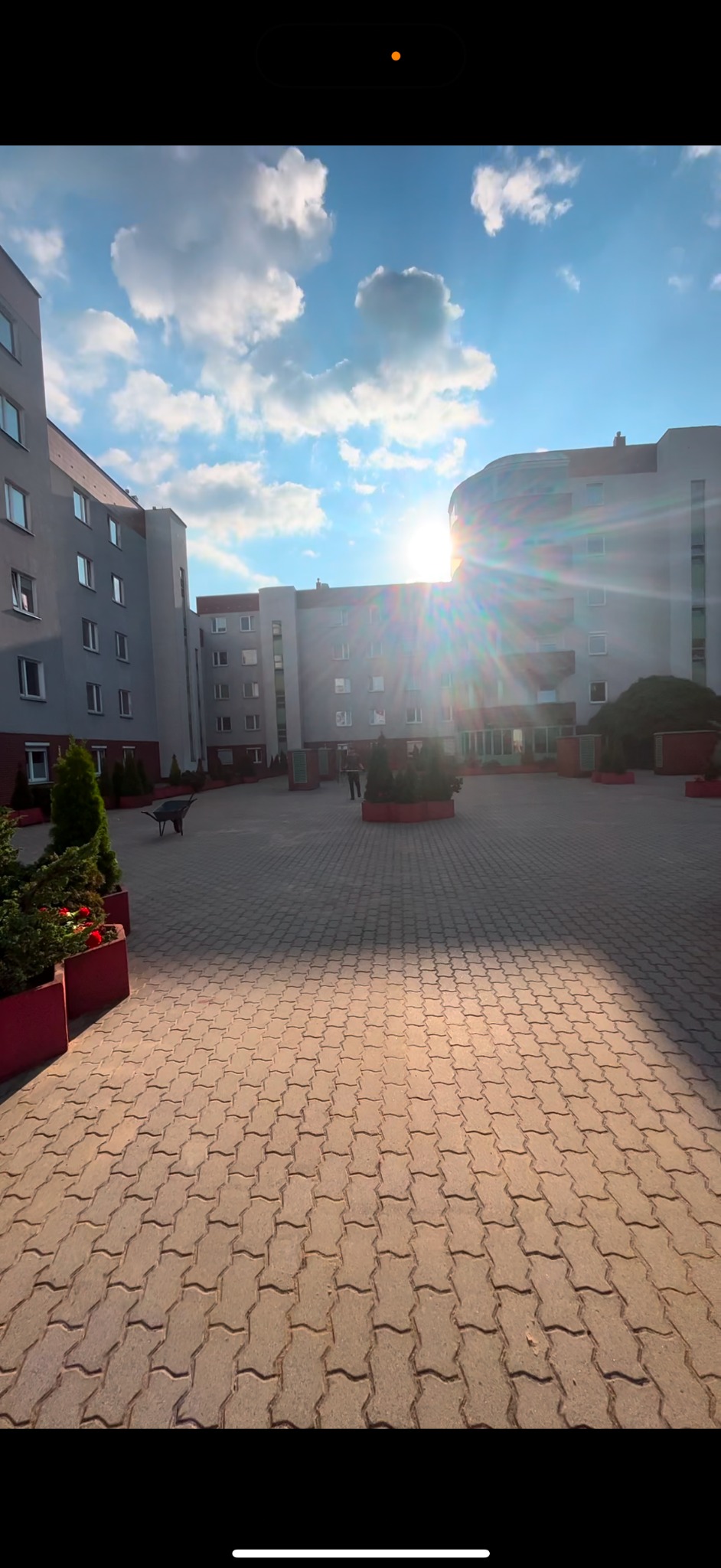 Dziedziniec wyłożony kostką brukową przed budynkiem mieszkalnym w słoneczny dzień, widoczne donice z zielenią i oddalona sylwetka osoby oraz wózek.