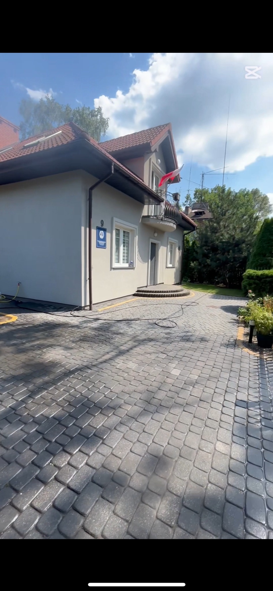 Mycie kostki brukowej na podjeździe przed budynkiem z polską flagą na balkonie, widoczne mokre ślady po czyszczeniu.