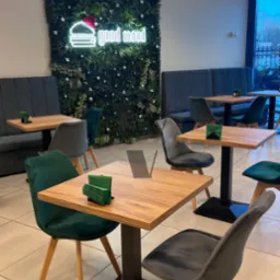 Wnętrze restauracji z drewnianymi stolikami na czarnych podstawach, krzesłami obitymi welurem w odcieniach szarości i zieleni oraz ścianą z roślin i neonowym napisem 'good mood' w tle.