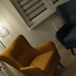 Pokój z dwoma fotelami: musztardowym i granatowym, oświetlony lampą podłogową z metalowym kloszem, z widokiem na okno z żaluzjami.