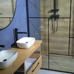 Nowoczesna łazienka z dwiema umywalkami nablatowymi, drewnianą szafką z wysuwanymi szufladami, kabiną prysznicową typu walk-in z czarnymi profilami i niebiesko-brązowymi płytkami na ścianach.