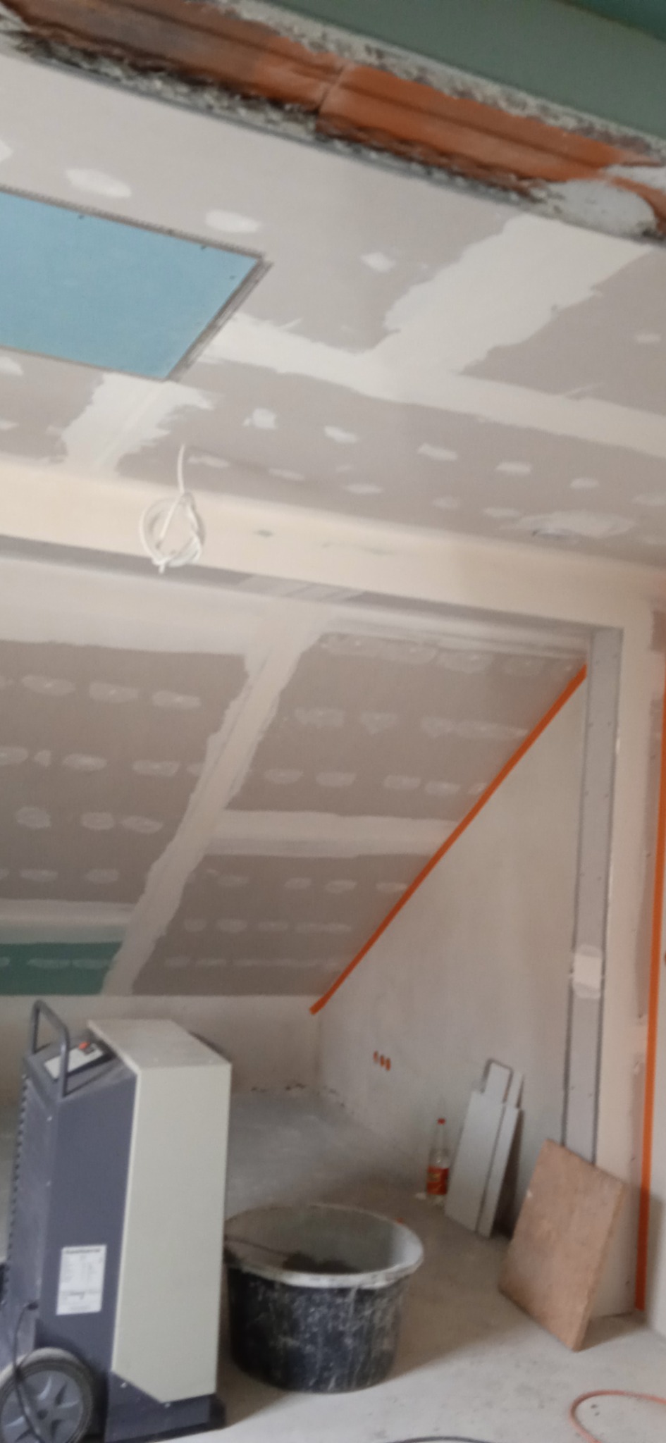 Skośne poddasze w trakcie prac wykończeniowych z zamontowaną płytą gipsowo-kartonową, widoczne fugi, taśma malarska, wiadro z zaprawą, osuszacz powietrza i otwór w suficie.