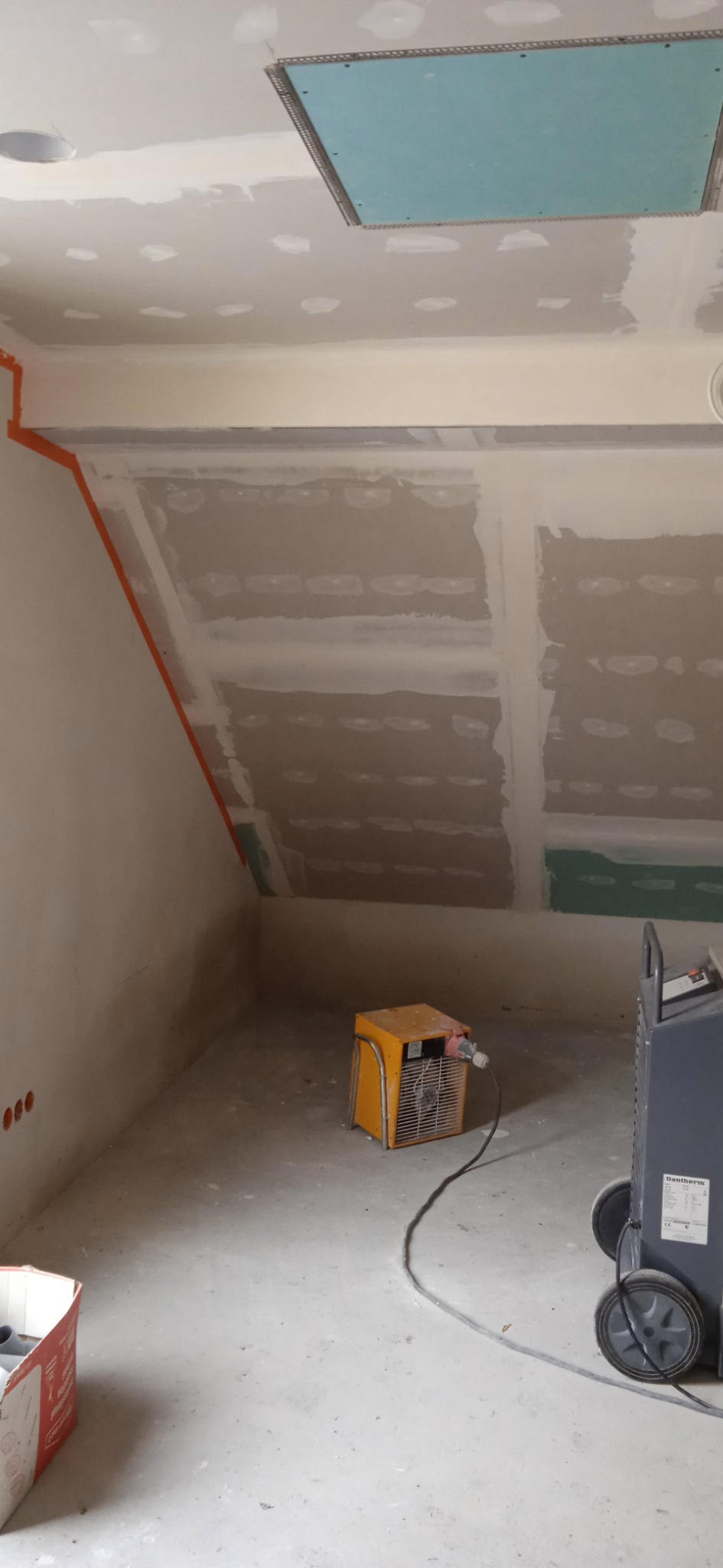 Poddasze w trakcie prac wykończeniowych: widoczne płyty kartonowo-gipsowe na skosach i suficie, szpachlowane łączenia, otwór rewizyjny w suficie, grzejnik elektryczny i osuszacz budowlany.