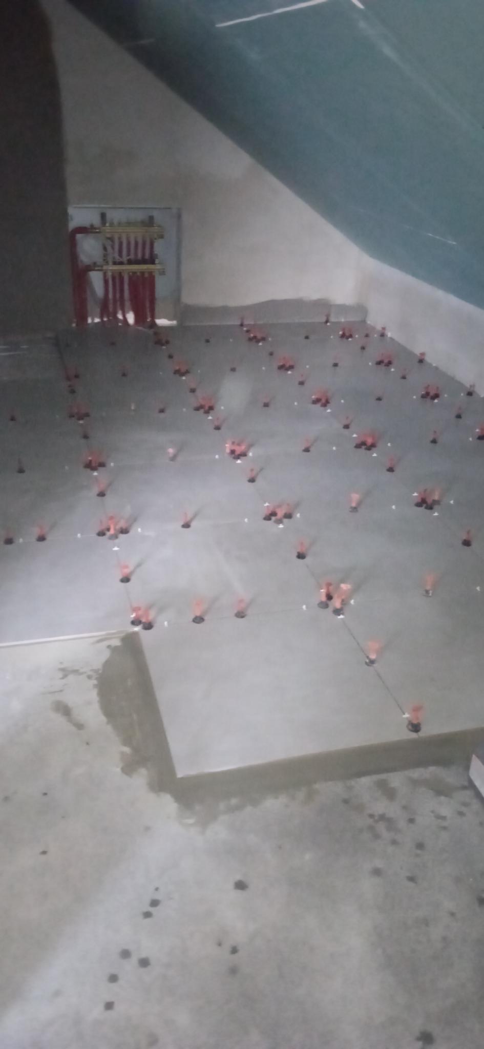 Układanie szarych płytek podłogowych w pomieszczeniu na poddaszu, widoczne czerwone kliny poziomujące, instalacja ogrzewania podłogowego na ścianie.