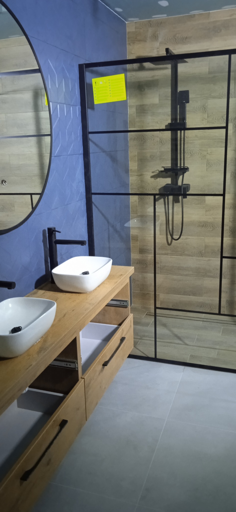 Nowoczesna łazienka z dwiema umywalkami nablatowymi, drewnianą szafką z wysuwanymi szufladami, kabiną prysznicową typu walk-in z czarnymi profilami i niebiesko-brązowymi płytkami na ścianach.